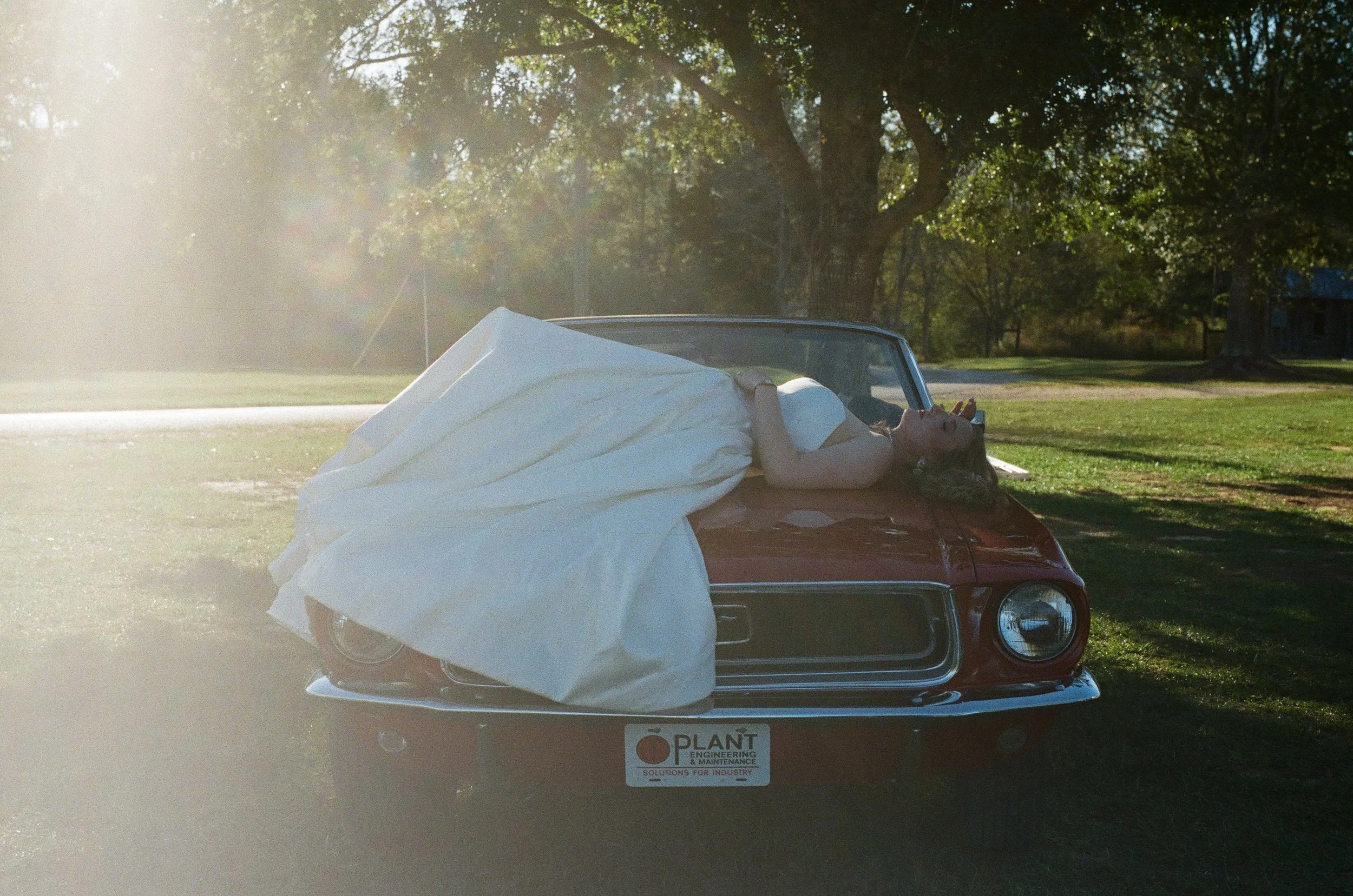 A woman in a white dress lying on the hood of a red vintage car, with her head back and eyes closed, in a grassy park with trees and sunlight shining through.