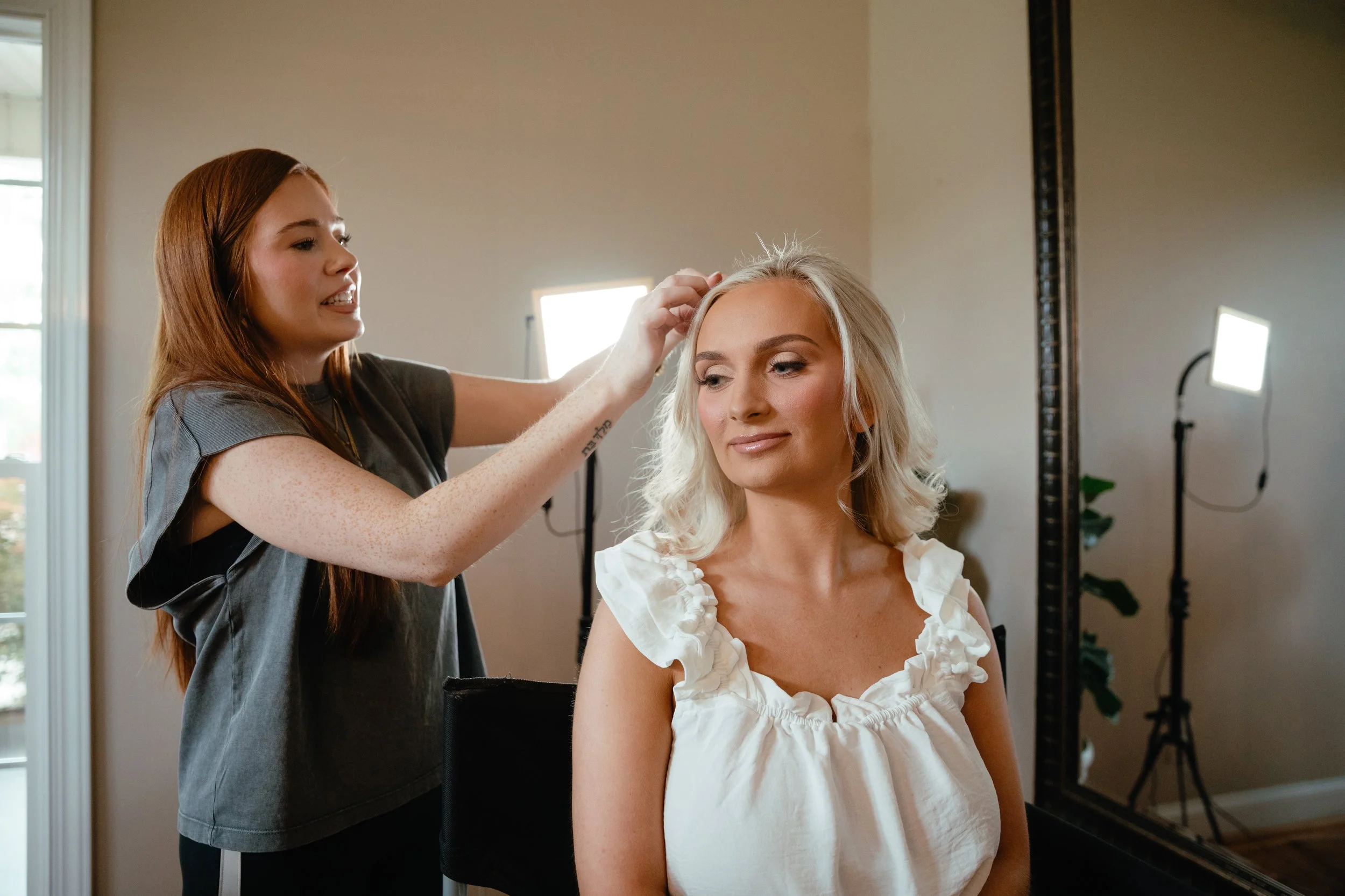 A woman with styled hair smiling while a stylist works on her hair in a decorated room.