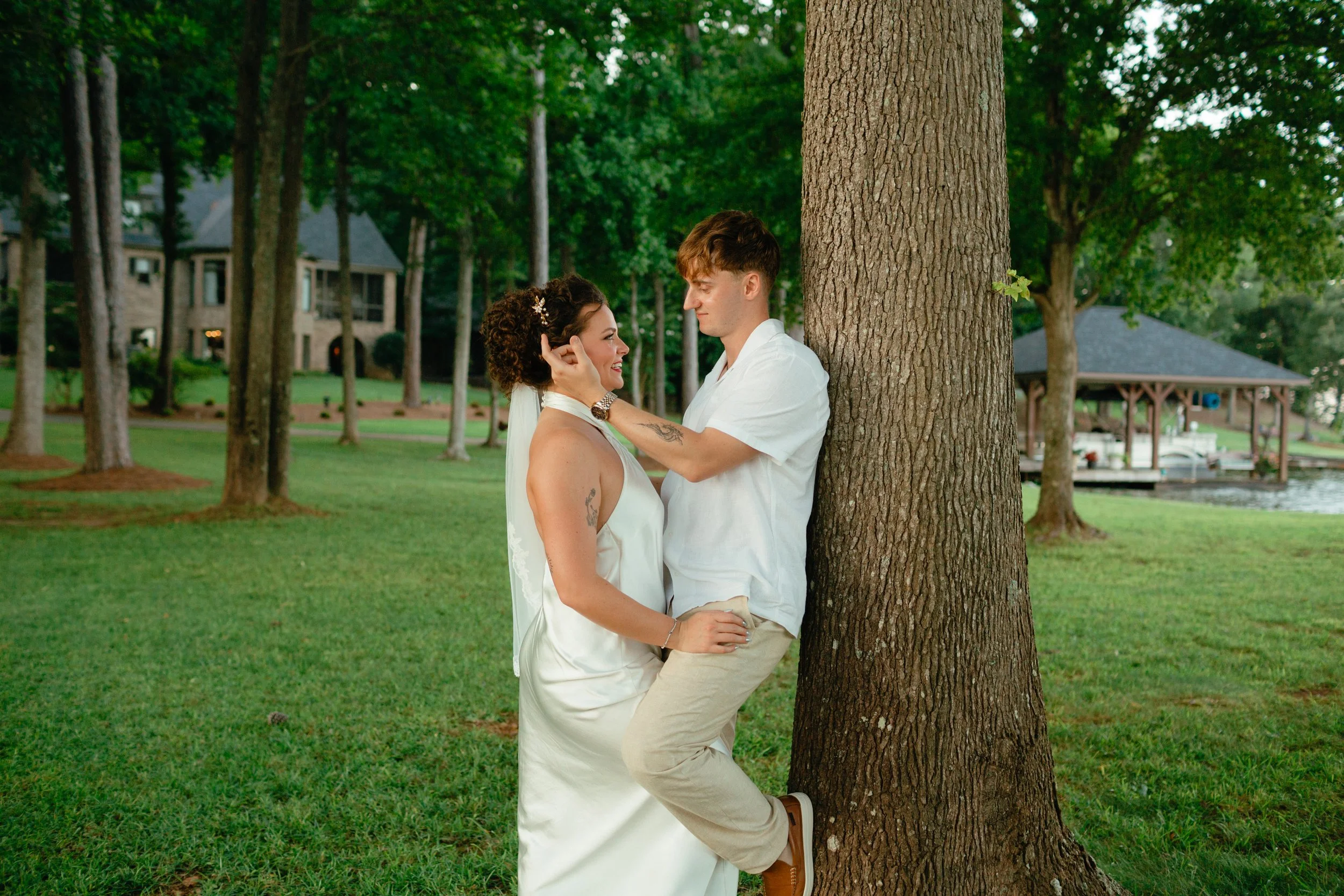 A couple standing close to each other in a park, with the woman touching the man's face and both smiling, near a tree with a grassy background and houses in the distance.