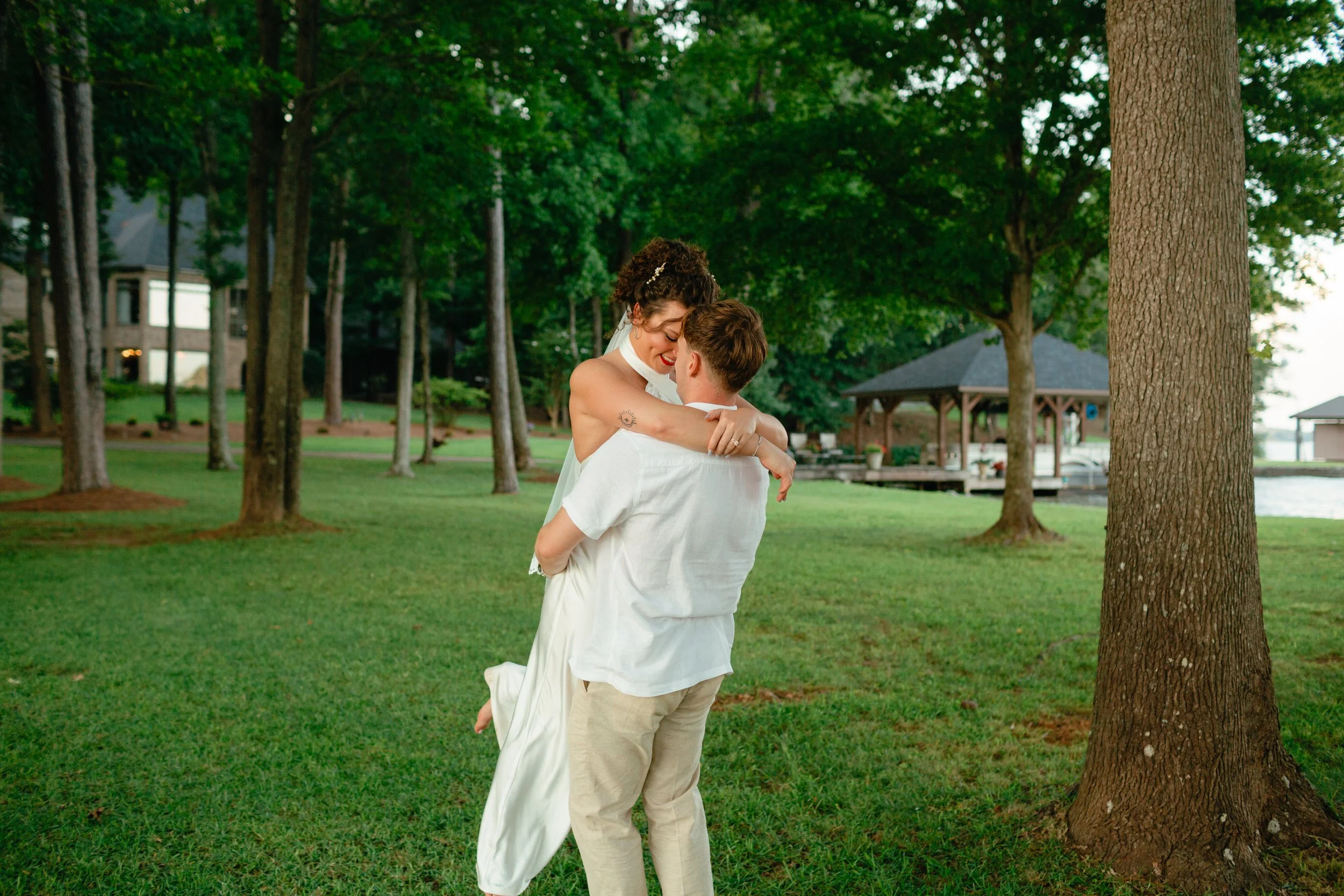 A couple embraces in a park with green grass, tall trees, and a gazebo in the background during daytime.