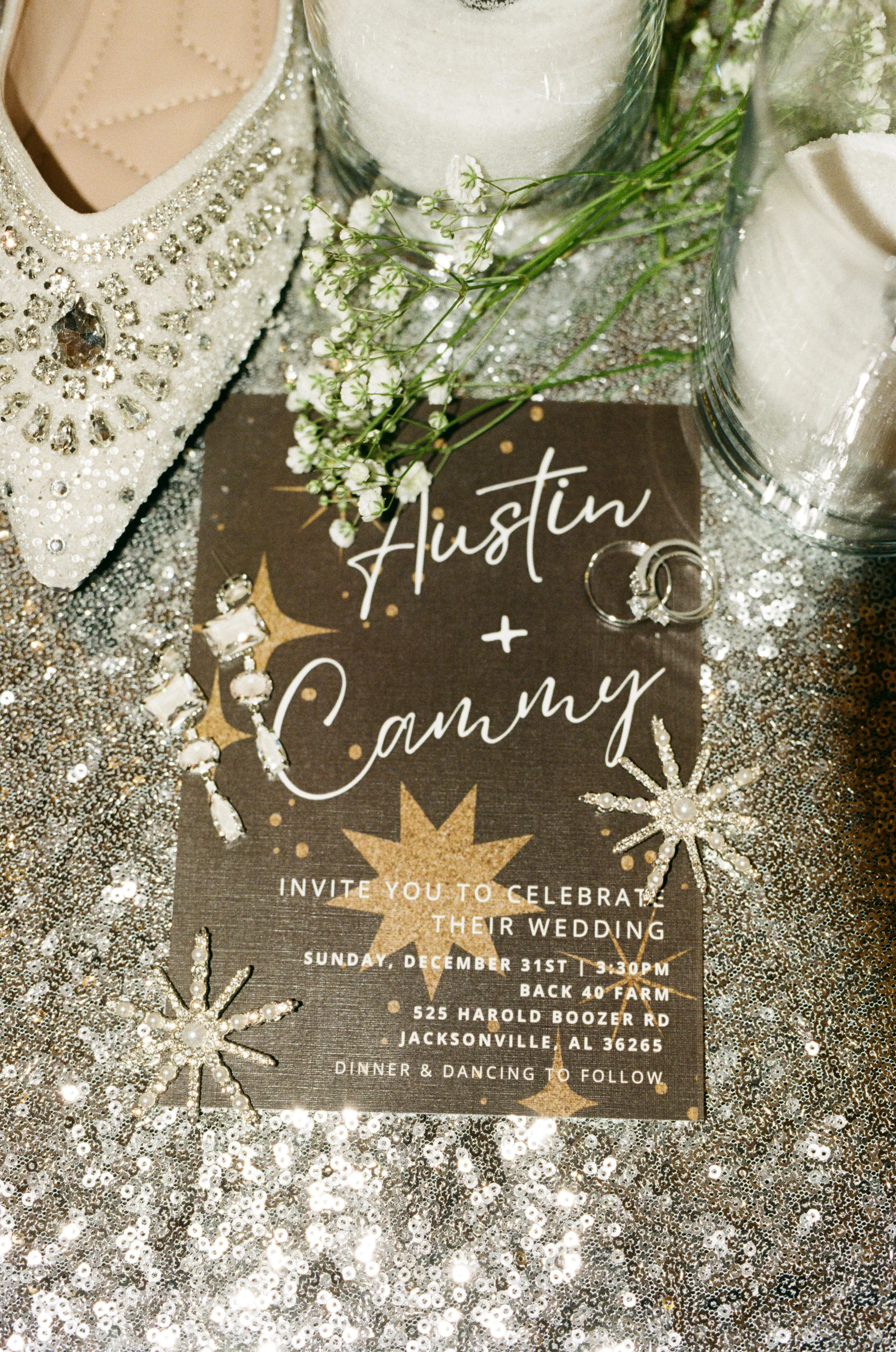 Wedding invitation for Austin and Cammy on a sparkly silver table, surrounded by jewelry, flowers, and candles. Back 40 farm in jacksonville, alabama, riley green's venue, 35mm film