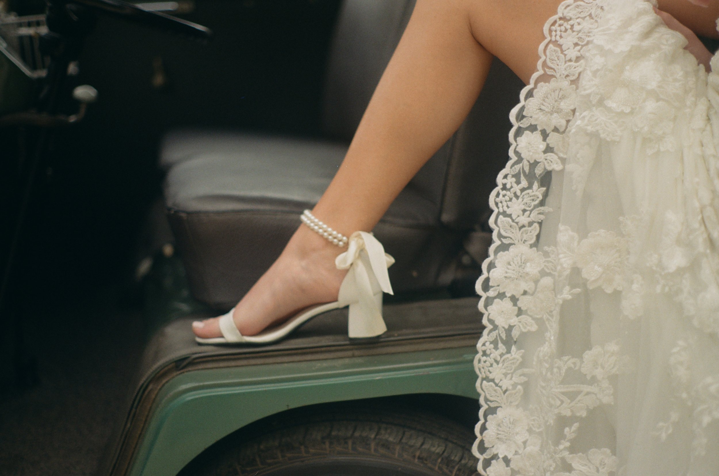 Close-up of a woman's hand with a pearl bracelet, resting on a white high-heeled shoe with a bow, next to a wedding dress with lace embroidery.