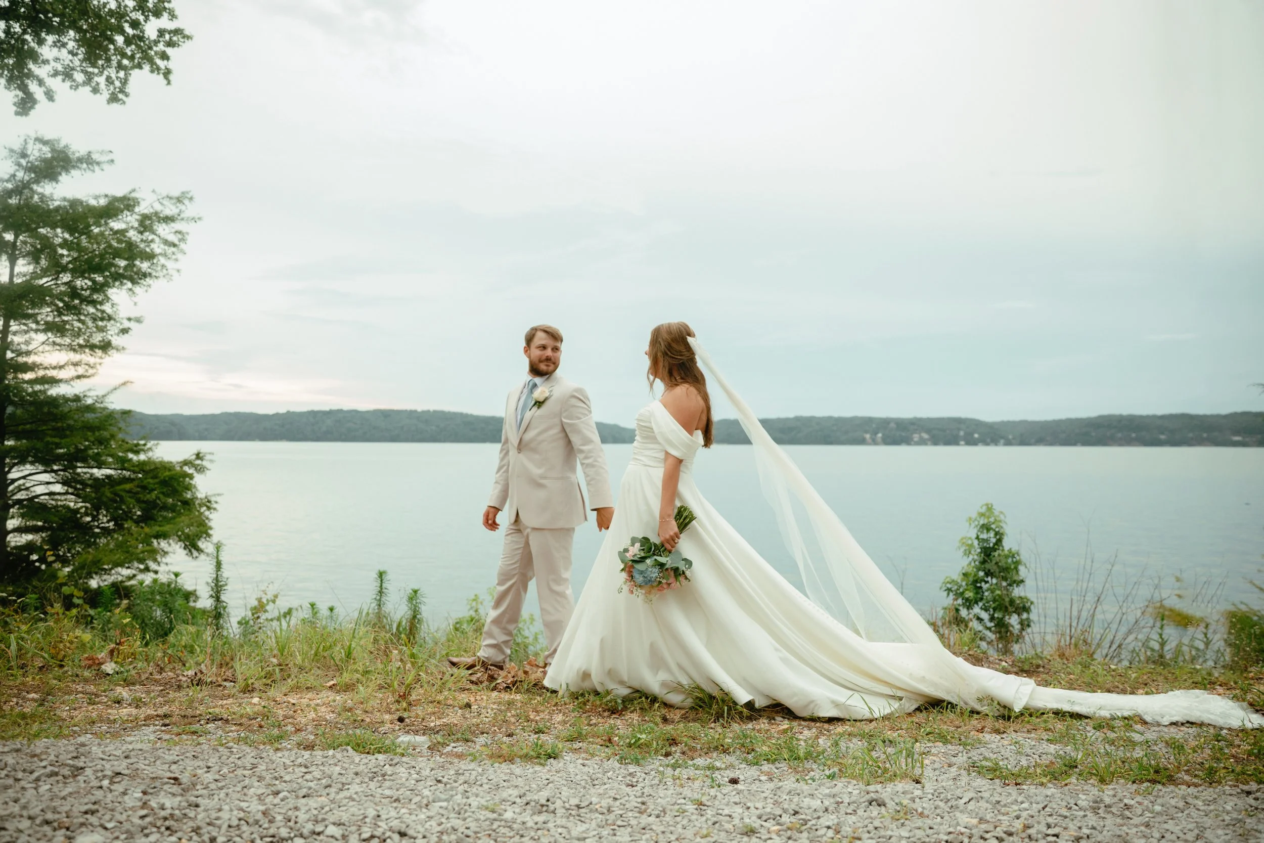 A bride and groom standing on a grassy area near a body of water, holding hands and looking at each other, during their outdoor wedding.