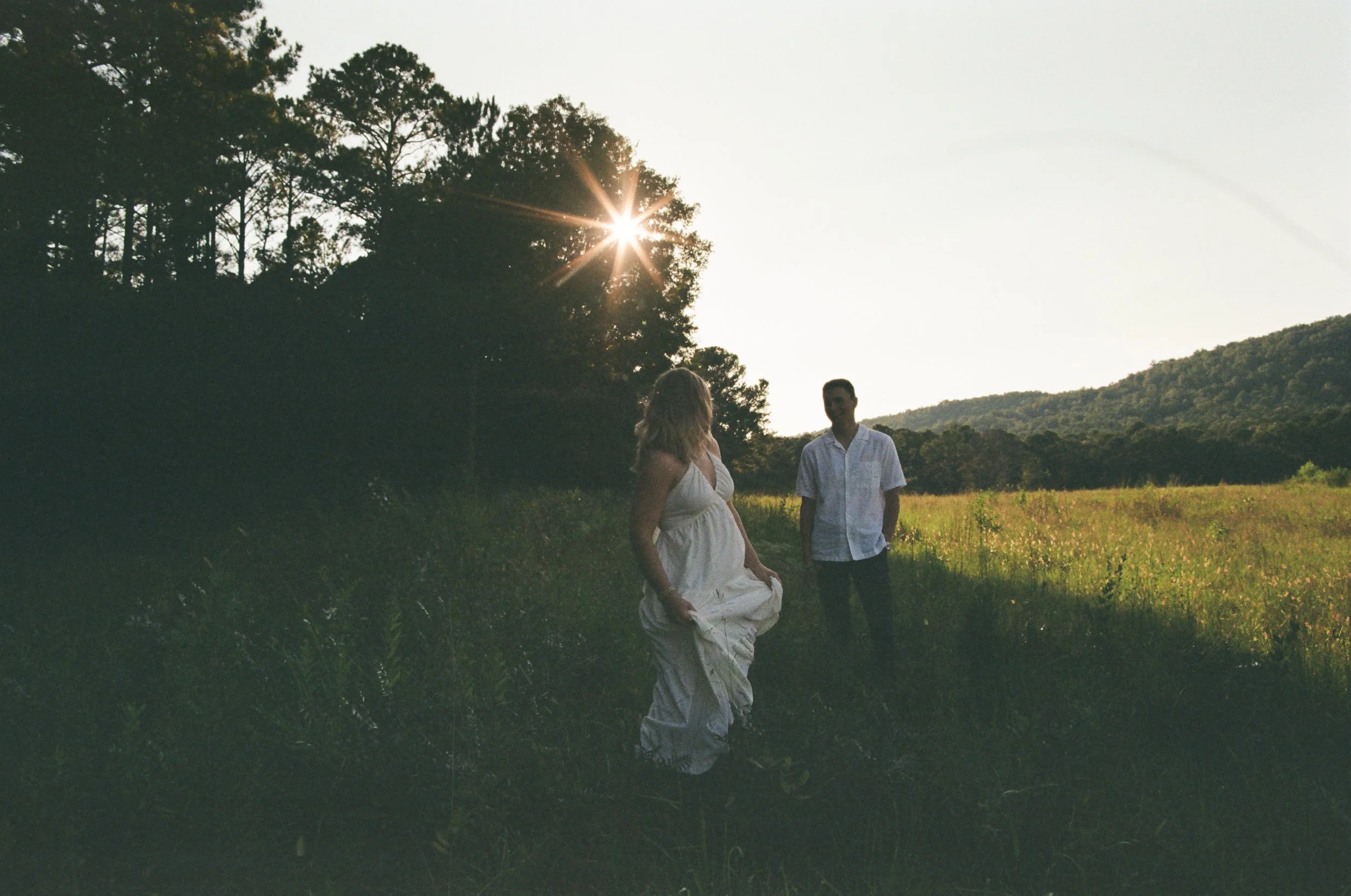 A woman in a white dress and a man in a white shirt standing outdoors in a grassy field with trees and mountains in the background, during sunset.
