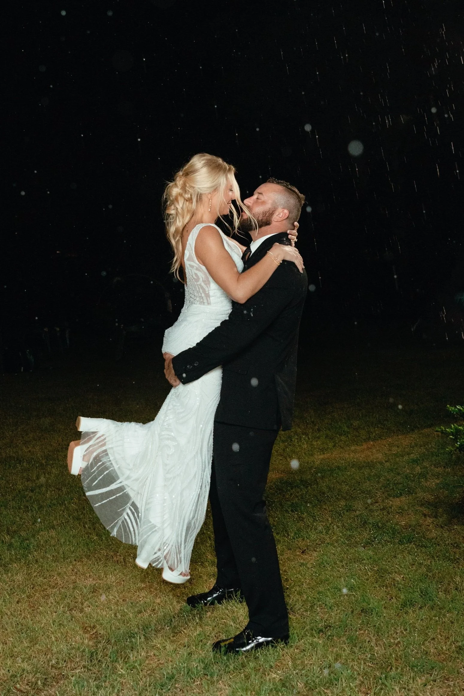 A newlywed couple dancing outdoors at night in the rain, with the groom lifting the bride off the ground.