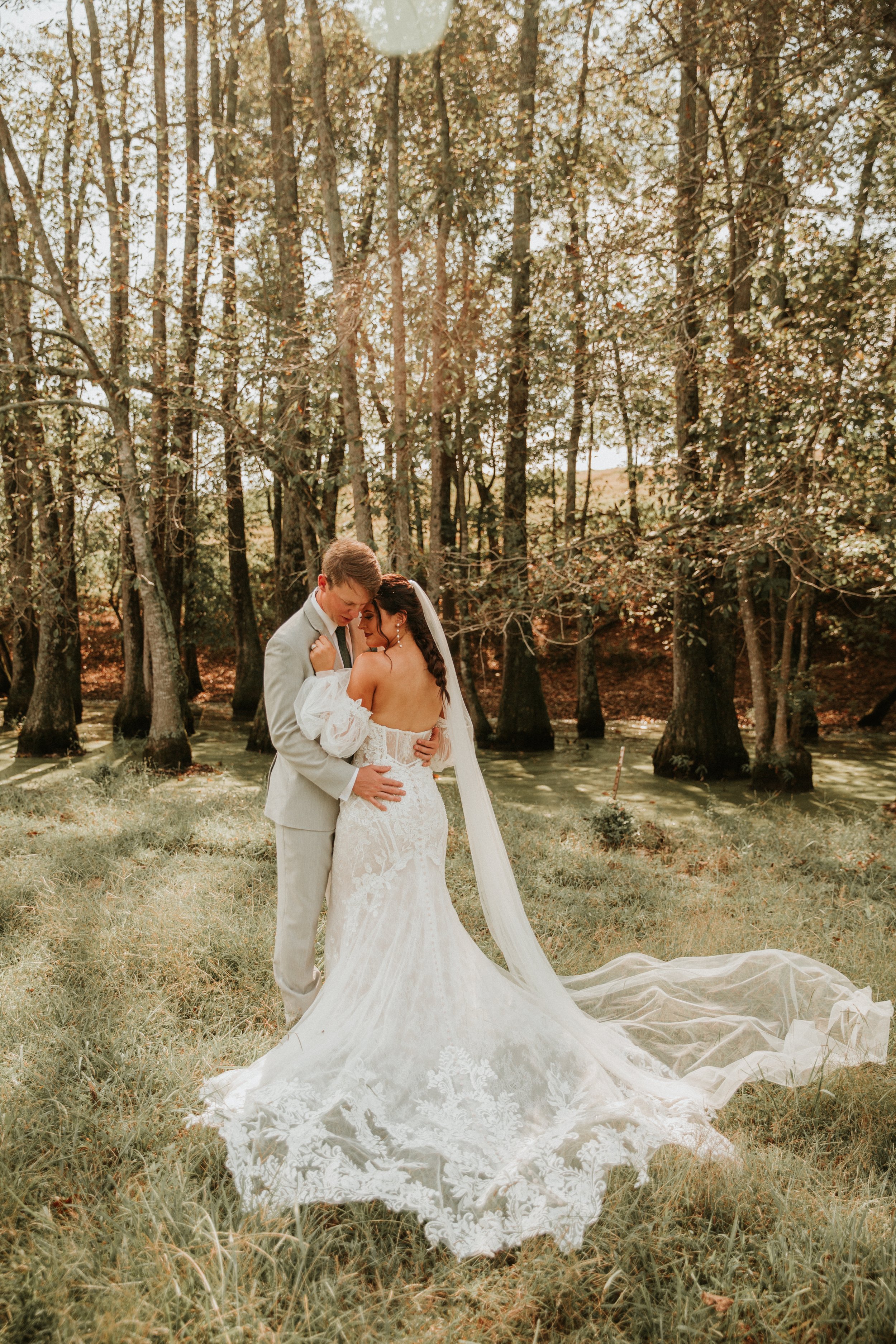 A bride and groom sharing a kiss outdoors next to a small creek in a wooded area during sunset, with the bride wearing a white lace wedding gown and long veil, and the groom in a light-colored suit.