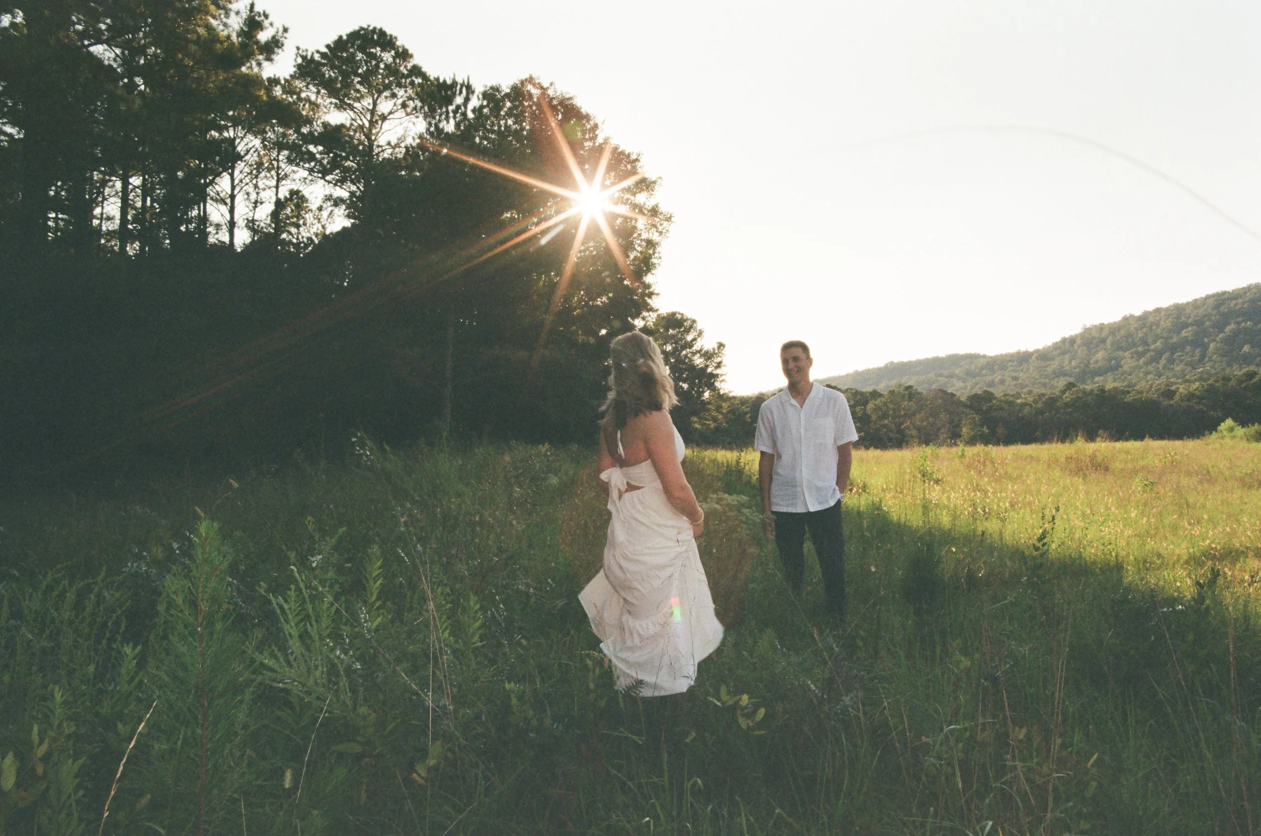 A couple standing in a grassy field during sunset, facing each other and smiling, with trees and hills in the background. Ridge Pointe on 35 mm film
