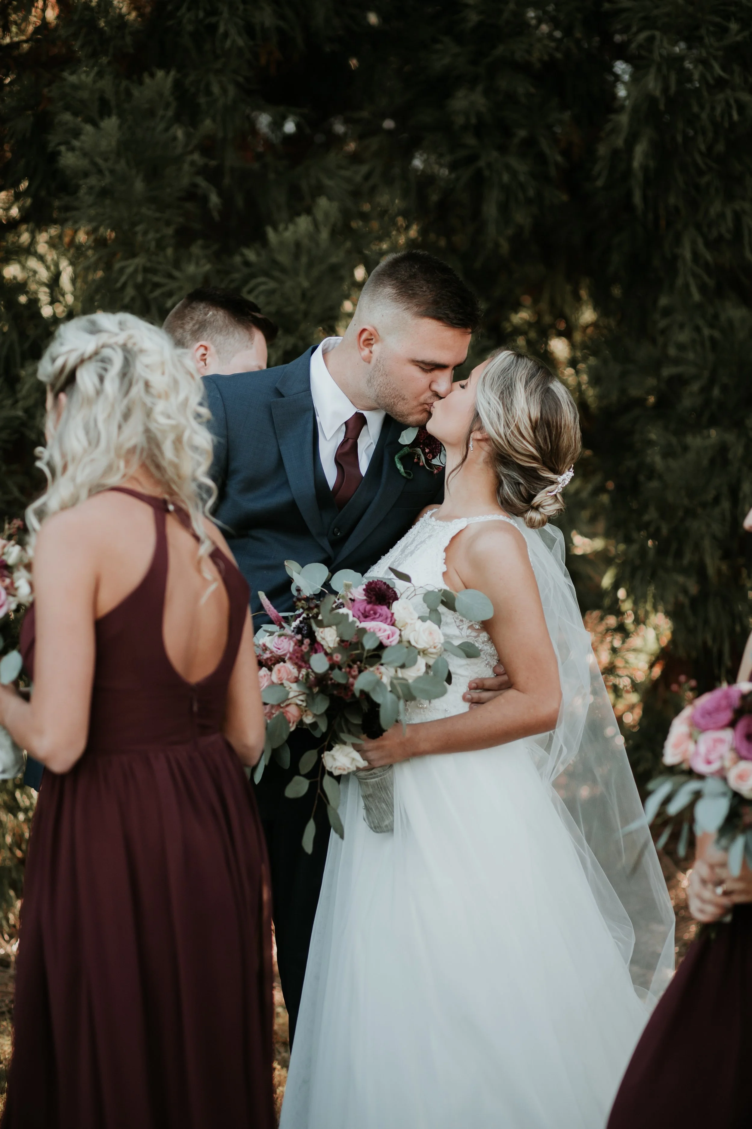 A bride and groom share a kiss during their wedding ceremony outdoors, surrounded by bridesmaids holding bouquets, with lush green trees in the background.
