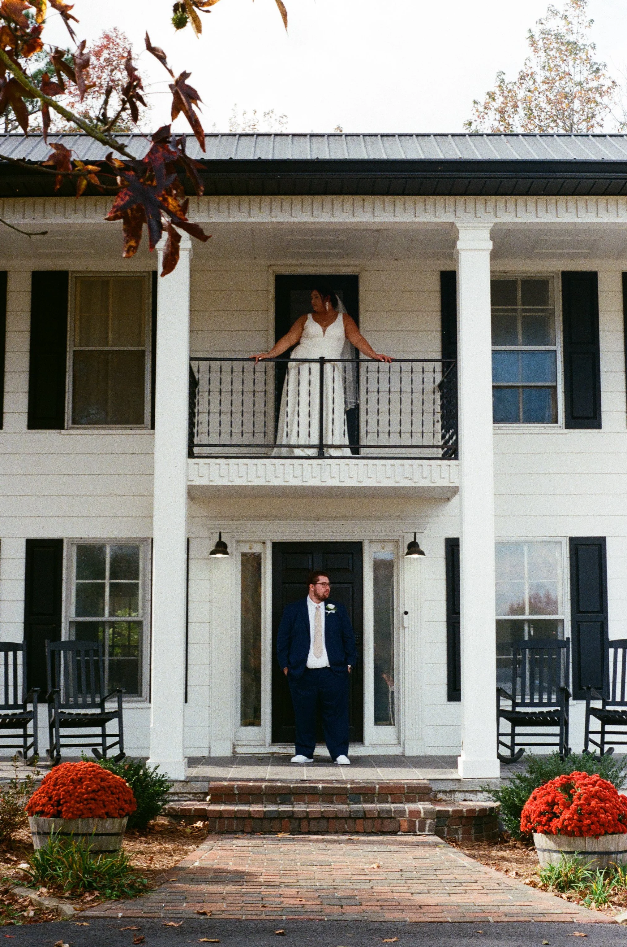 A bride in a white wedding dress standing on a balcony above a groom in a dark suit standing at the front door of a white house with black shutters, surrounded by potted orange flowers on 35mm film