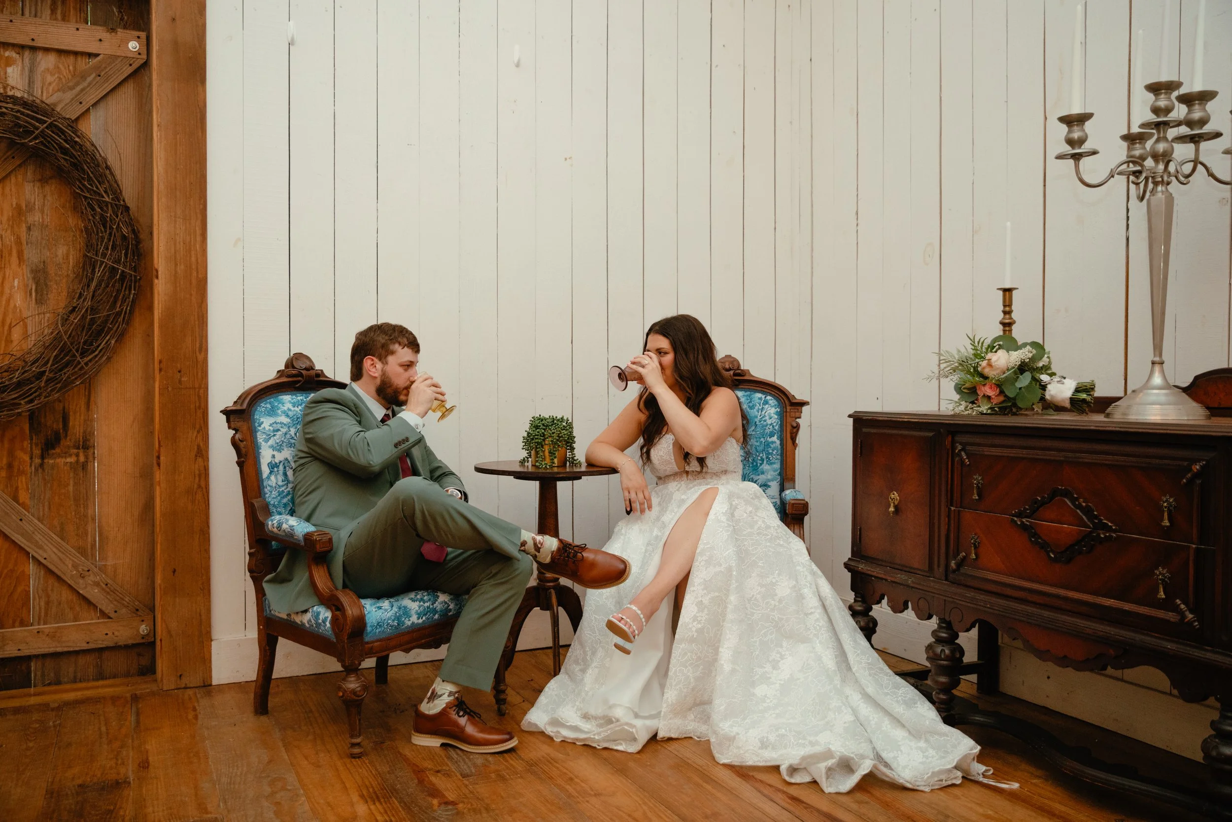 A bride and groom sitting on vintage chairs indoors, drinking from glasses, with a white wall, wooden furniture, a candelabra, and a floral arrangement nearby.