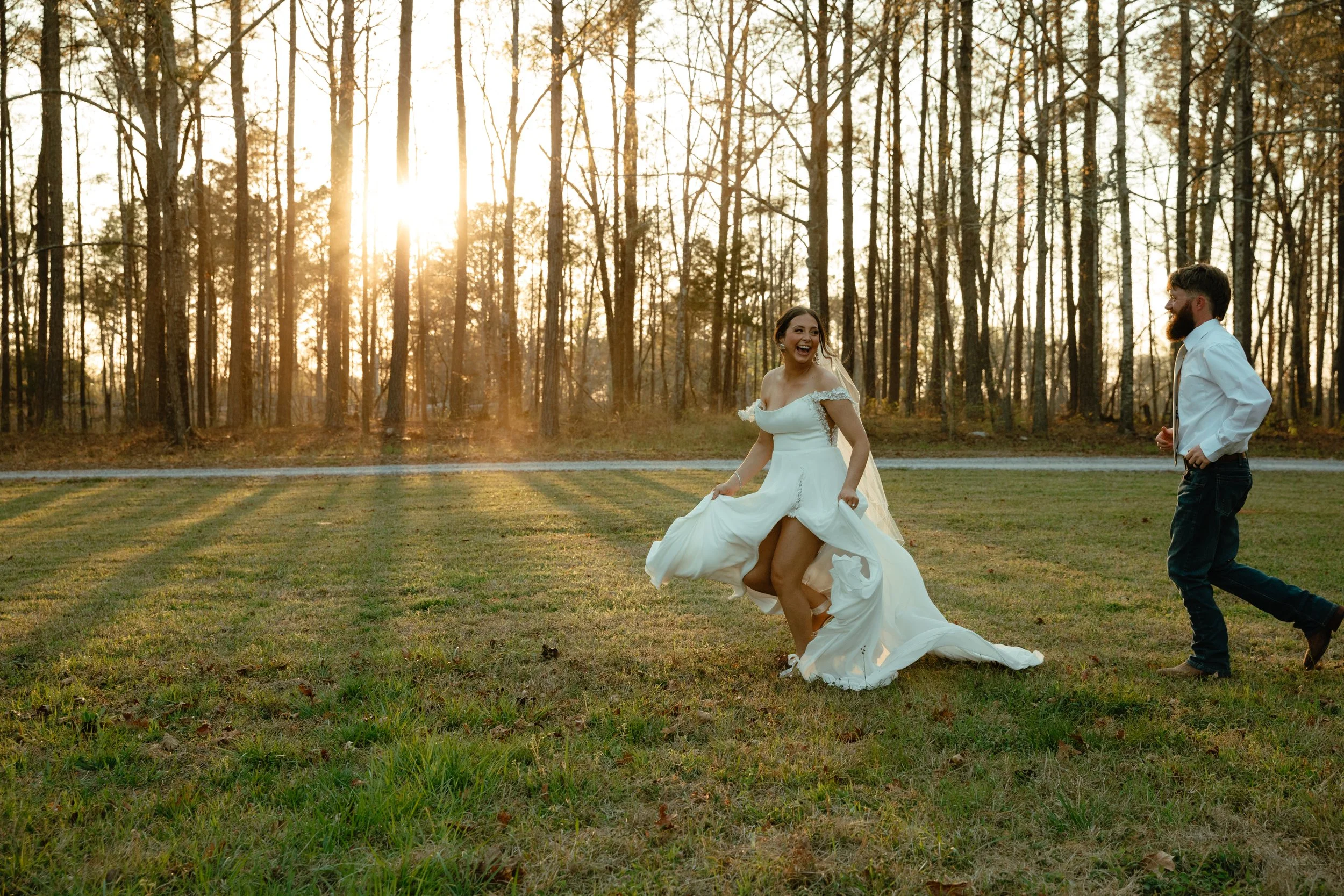 A bride in a white wedding dress dancing on a field with trees in the background during sunset, smiling and laughing while a groom in casual attire watches her.
