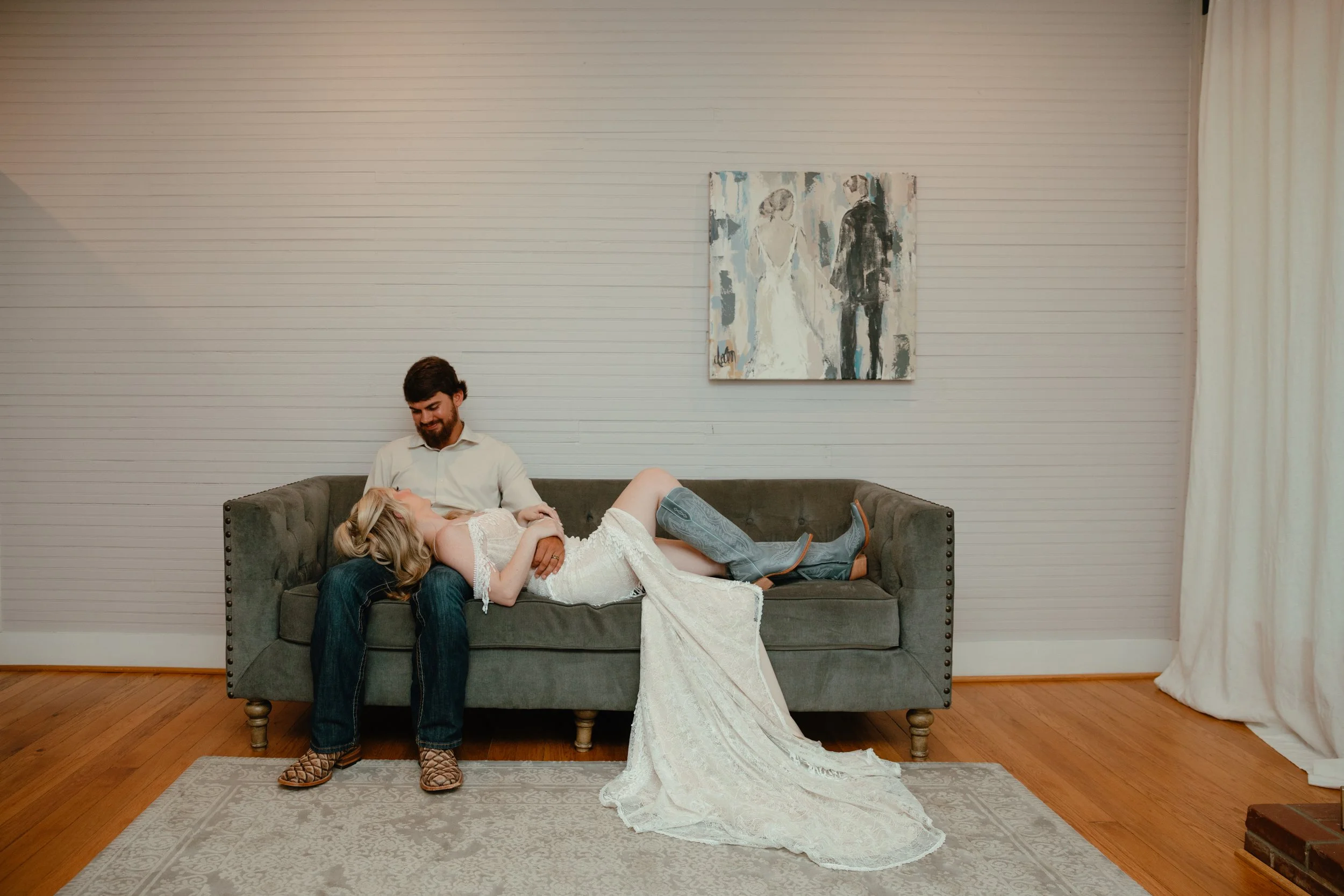 A couple sitting on a sofa with a woman lying on the man's lap, wearing a wedding dress, in a room with white paneled walls, a painting, and curtains on the right, with wooden flooring and a gray rug.
