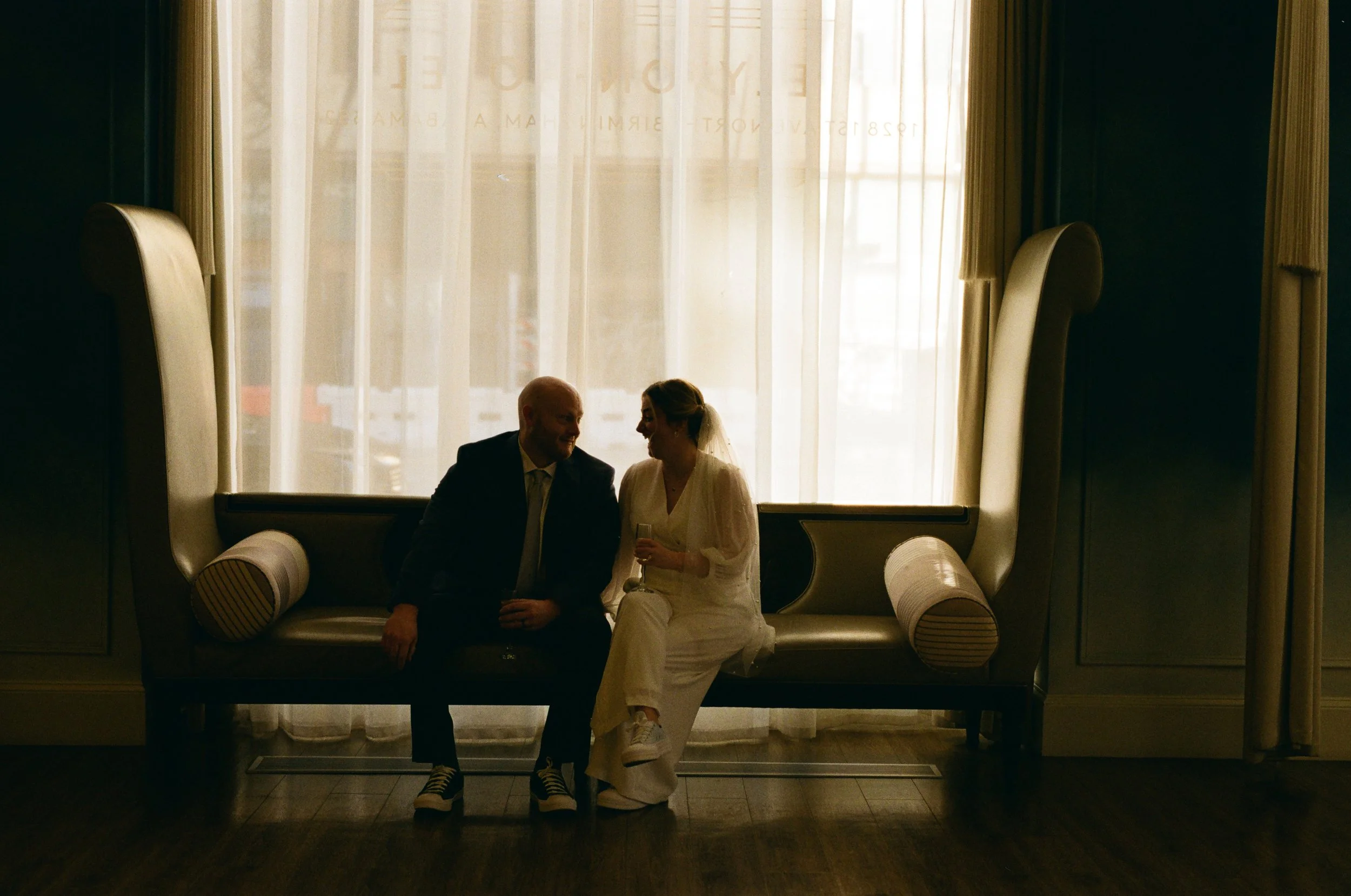 Silhouetted couple sitting on a bench in front of large window, smiling and sharing a moment, with the woman holding a glass of champagne. Elyon Hotel in birmingham, alabama on 35mm film