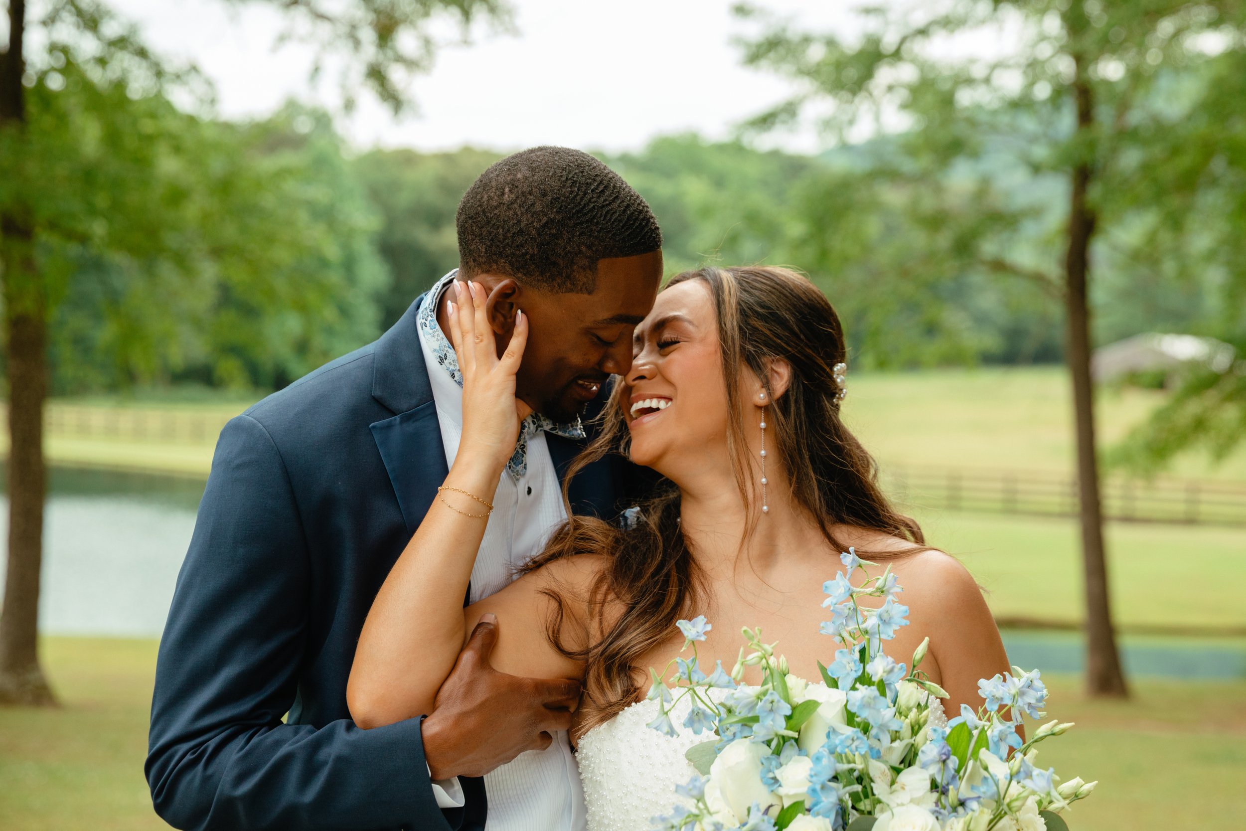 A newlywed couple sharing a joyful moment outdoors, the groom leaning into the bride with both smiling and eyes closed, surrounded by green trees and holding a bouquet of white and blue flowers.