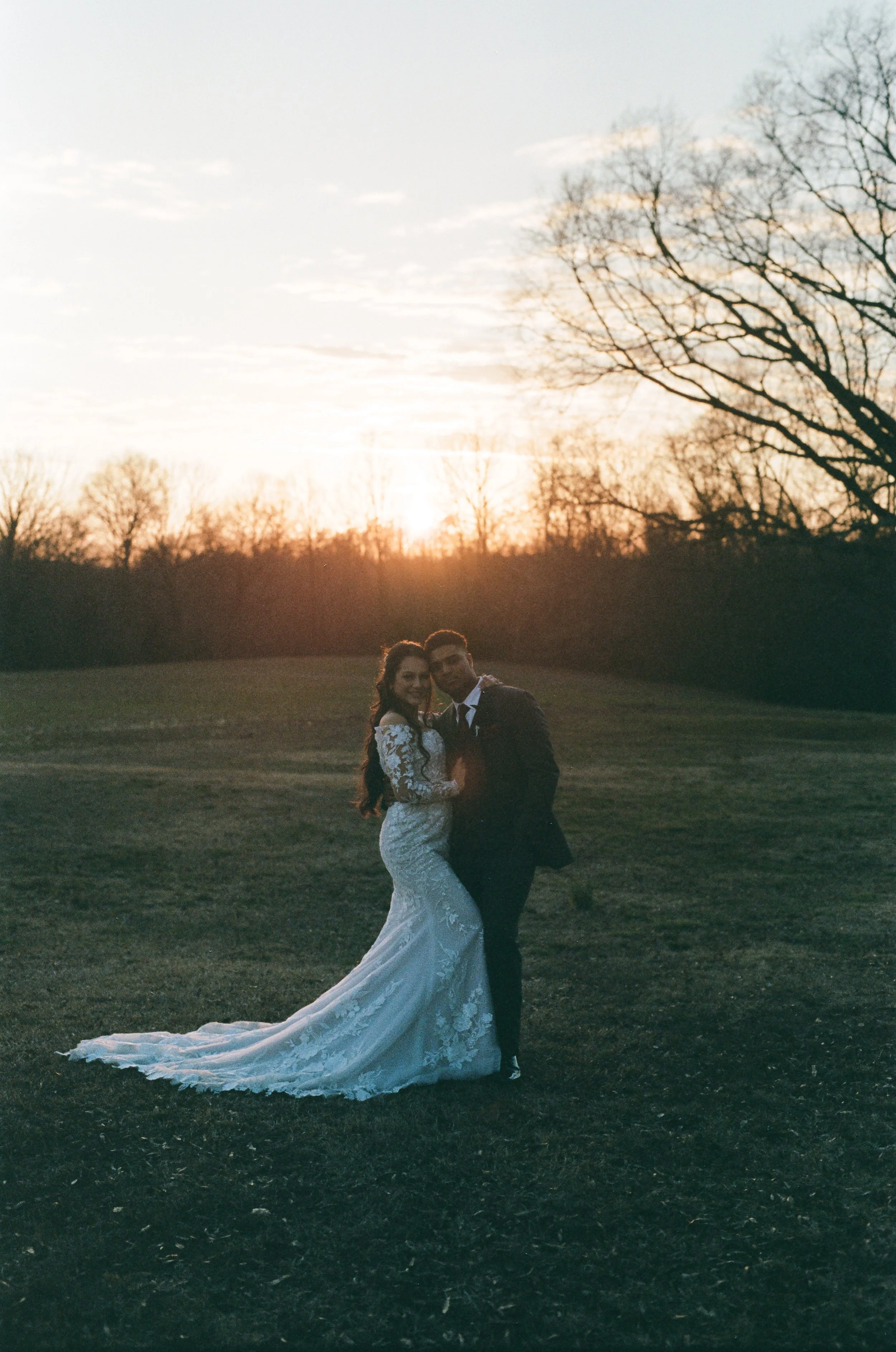 A bride and groom stand together on a grassy field at sunset, with leafless trees in the background.