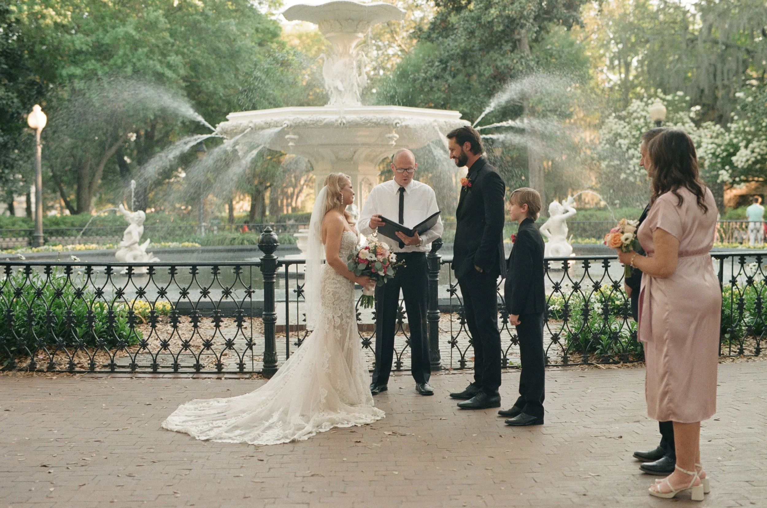 Wedding ceremony taking place outdoors near a fountain in a park, with a bride in a wedding dress holding a bouquet, a groom in a dark suit, officiant, bridesmaid, and a young boy, all standing on a brick pathway.