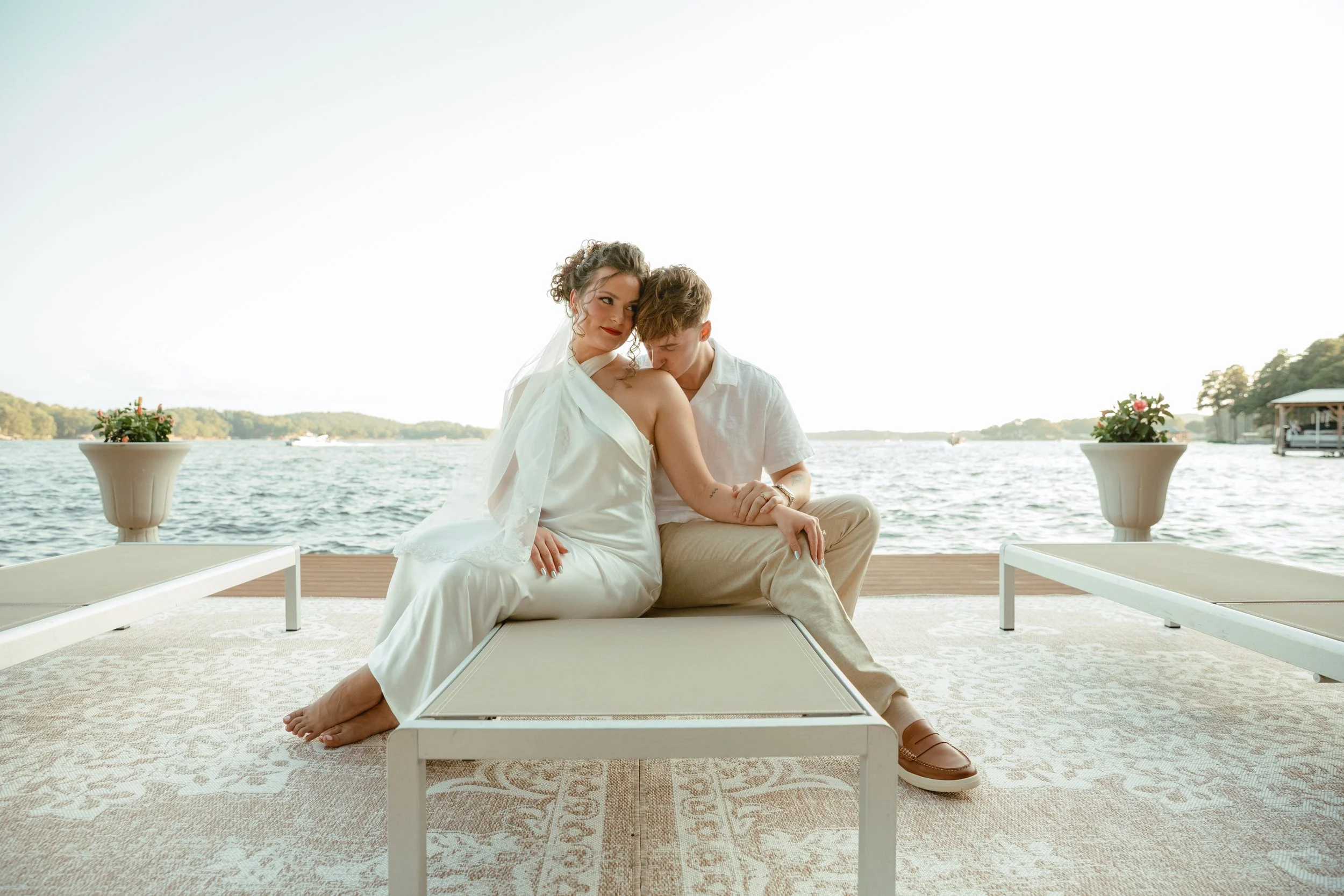 A couple sitting on a bench by the water at a lake or river with trees in the background, two potted plants with pink flowers on either side, the woman wearing a wedding dress and veil, the man in casual light-colored clothing, both embracing lovingl