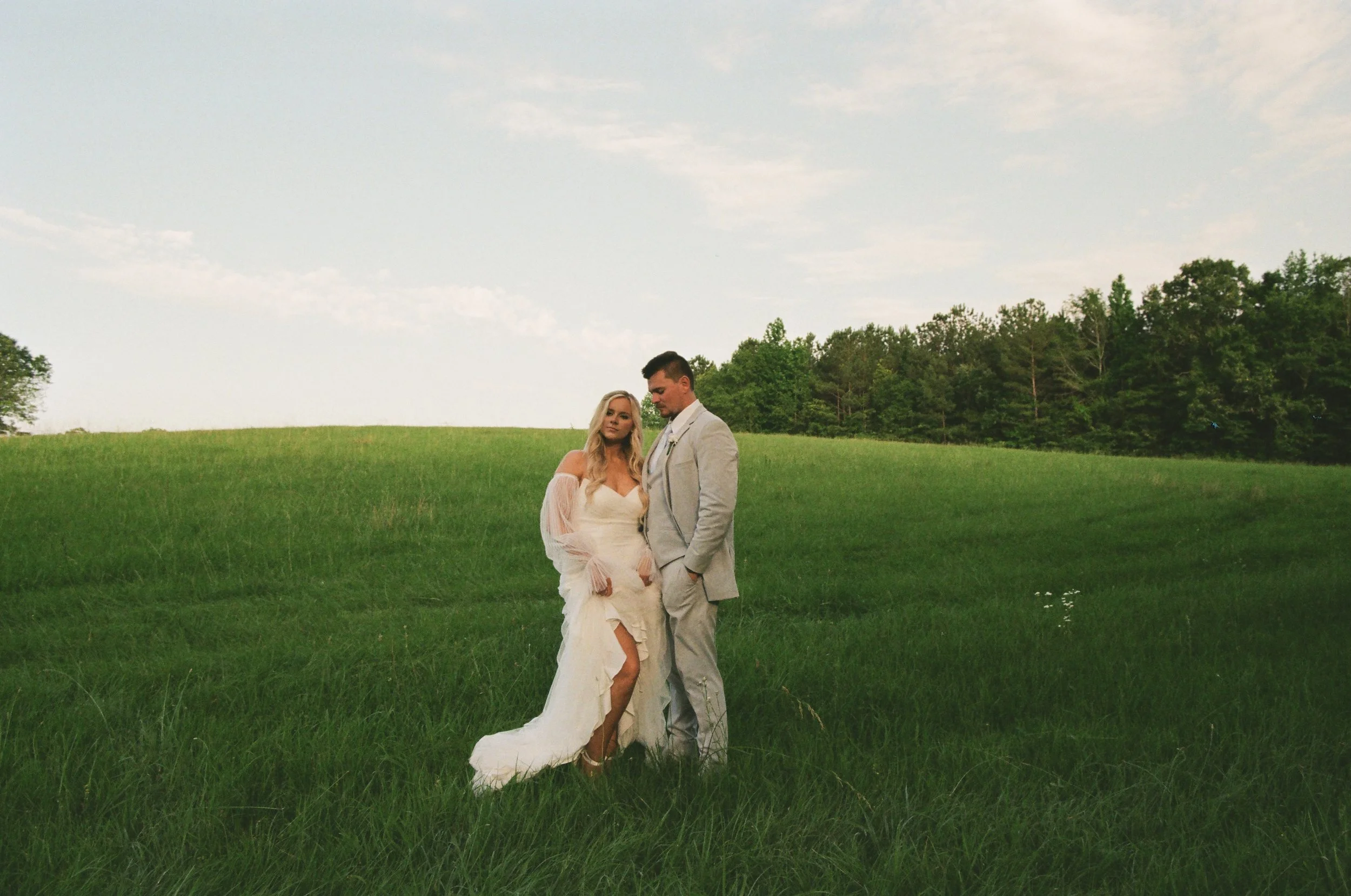A bride and groom standing in a green field under a sky with scattered clouds, dressed in wedding attire.