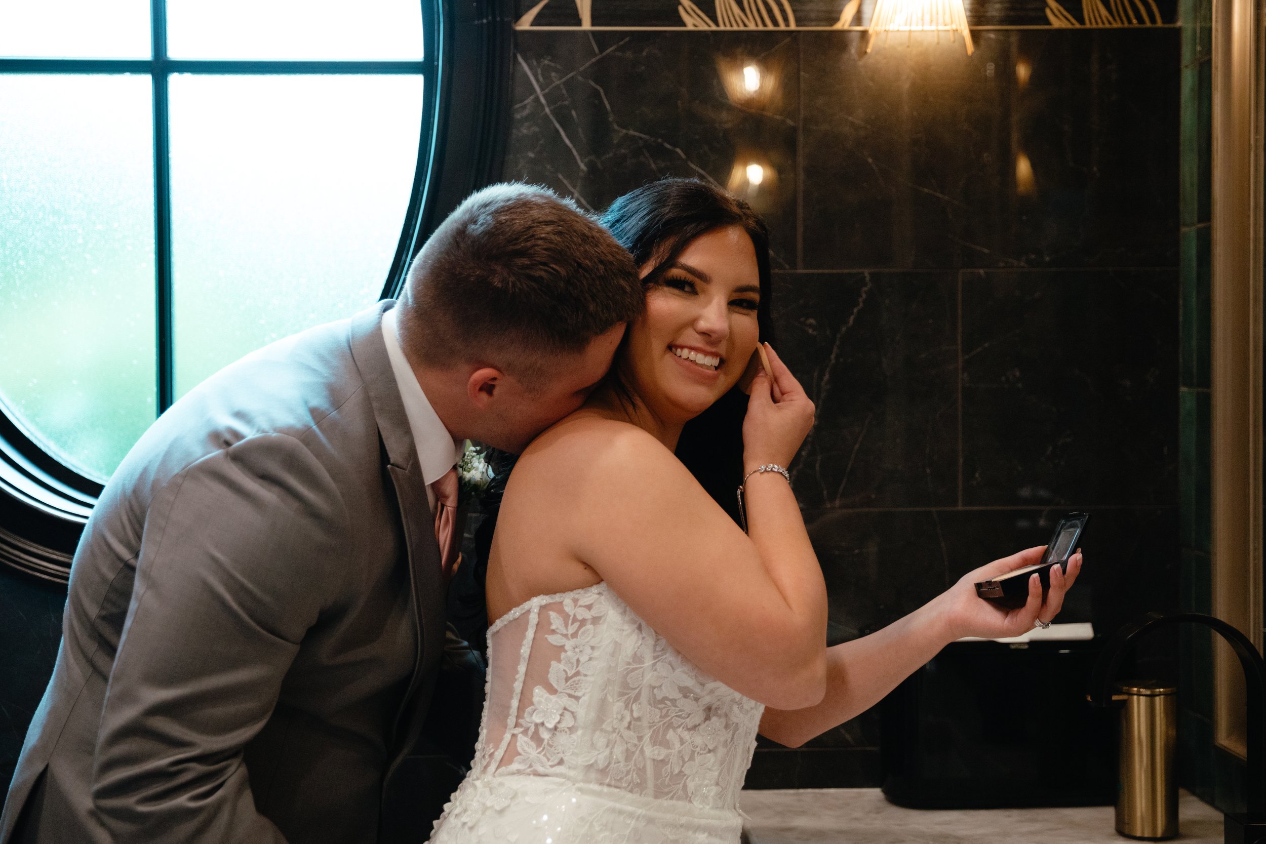 A bride in a white lace wedding dress smiling as a groom in a gray suit whispers into her ear, while she holds makeup in her hand, inside a dimly lit room with a large round window and black marble walls at The Lumpkin House, Birmingham