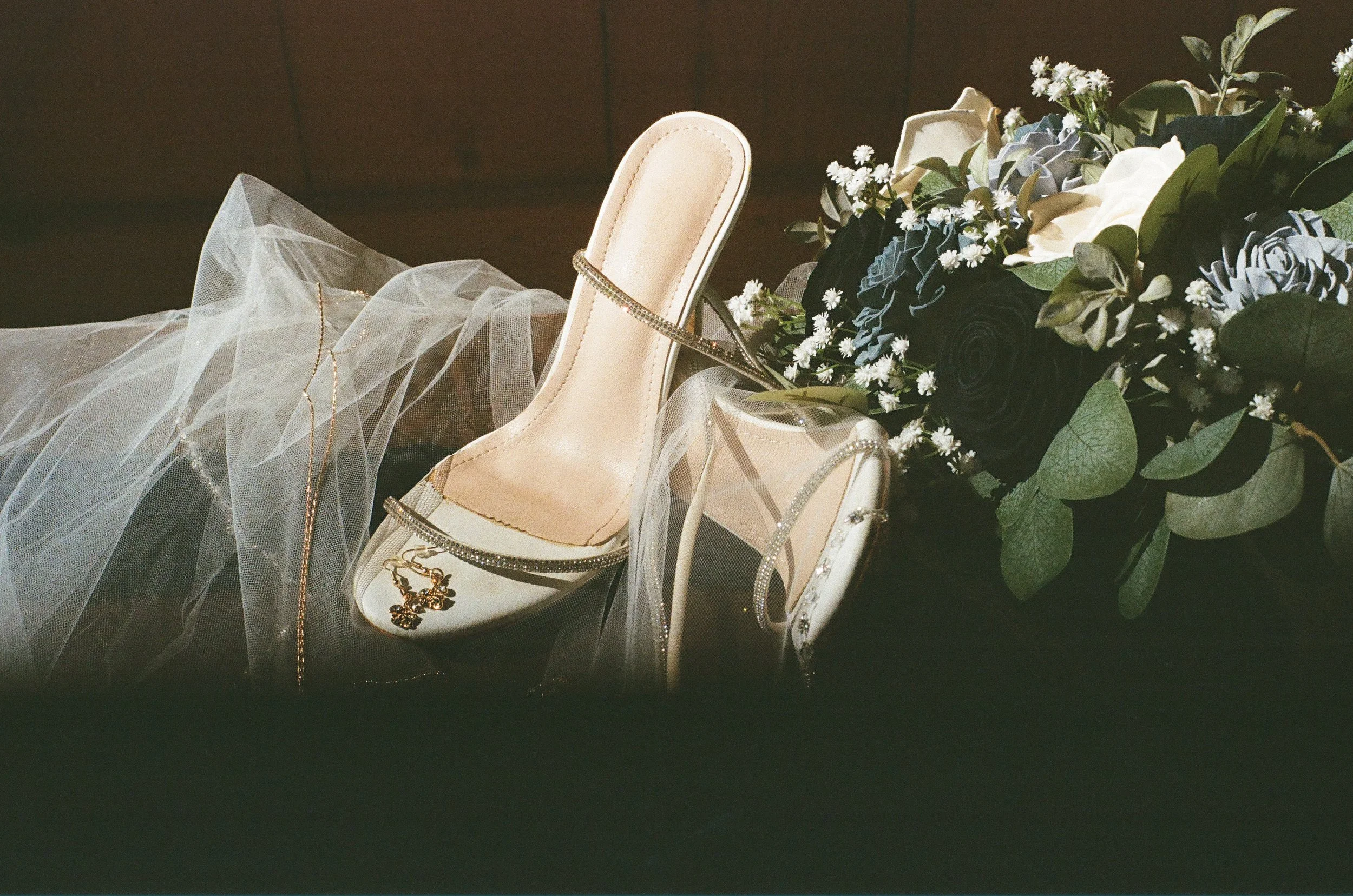 A pair of elegant white high-heeled shoes decorated with rhinestones, a cozy bouquet of black and white roses with greenery, and a sheer veil or fabric with gold jewelry, all arranged on a dark surface.
