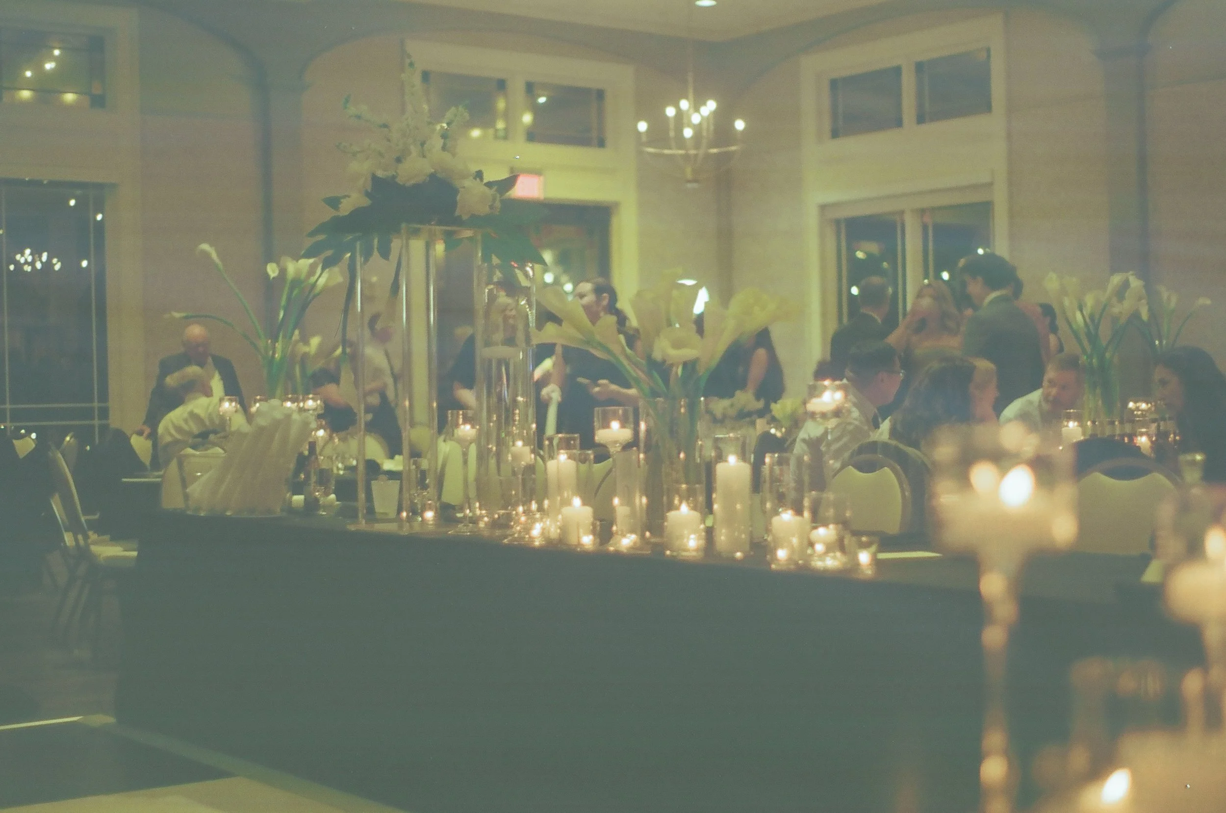 Elegant indoor banquet hall decorated with tall floral arrangements and numerous lit candles, with guests socializing in the background on 35mm film