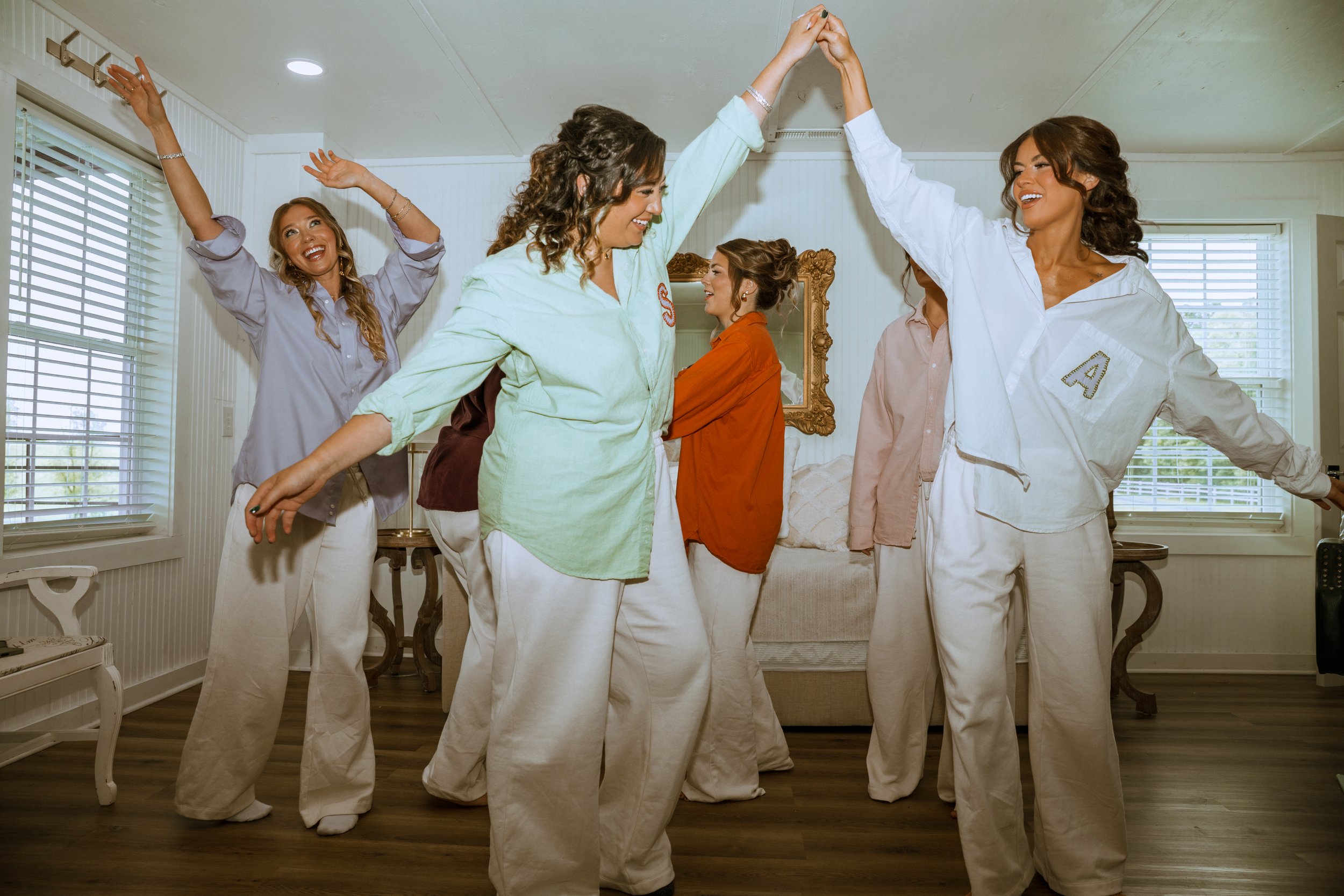 Group of five women indoors, wearing pajamas, high-fiving and smiling, with sunlight coming through window blinds.