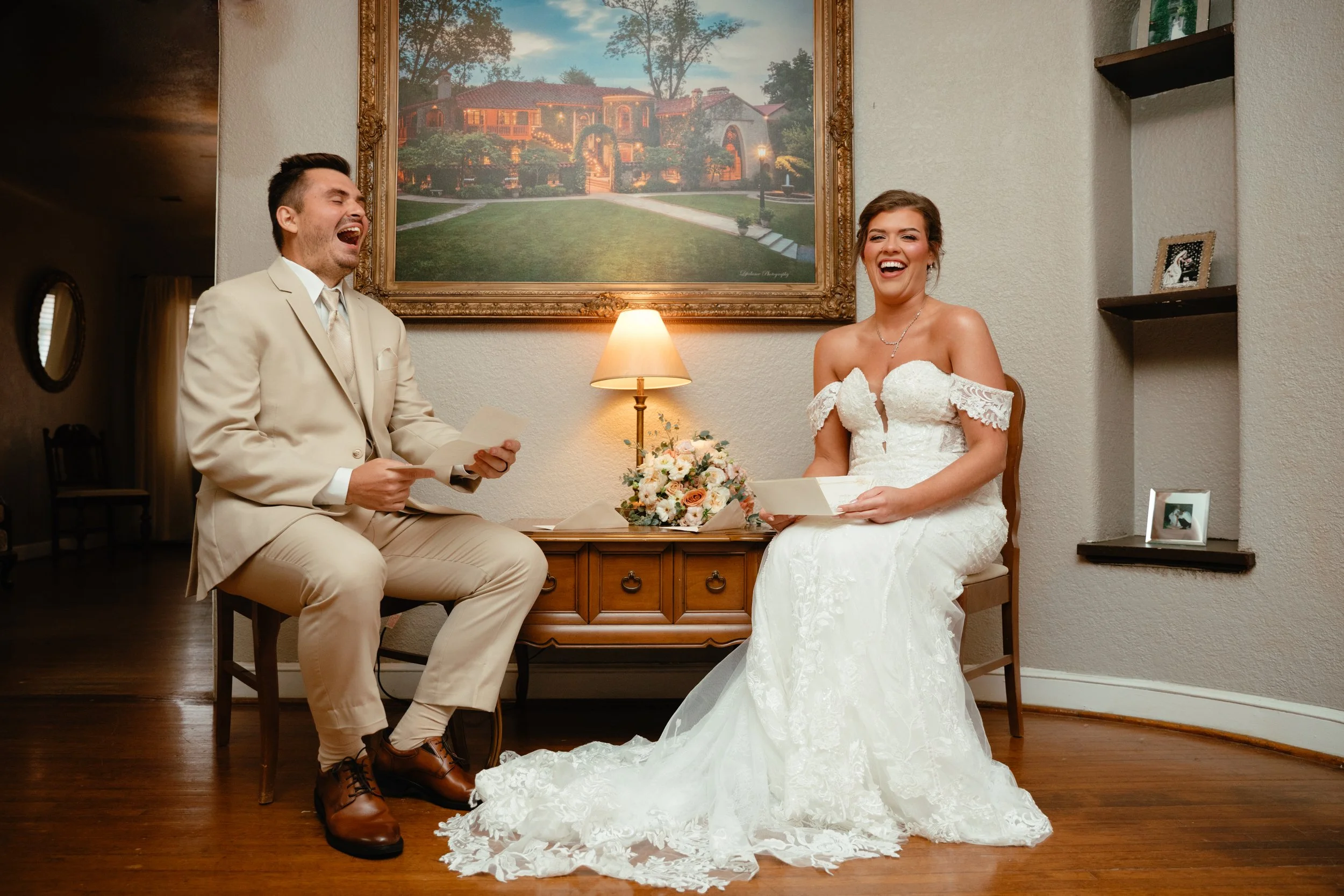 A bride and groom laughing during their wedding ceremony indoors, with a painting of a house and yard behind them. The groom wears a cream-colored suit and the bride wears a white lace wedding dress, holding a bouquet of flowers.