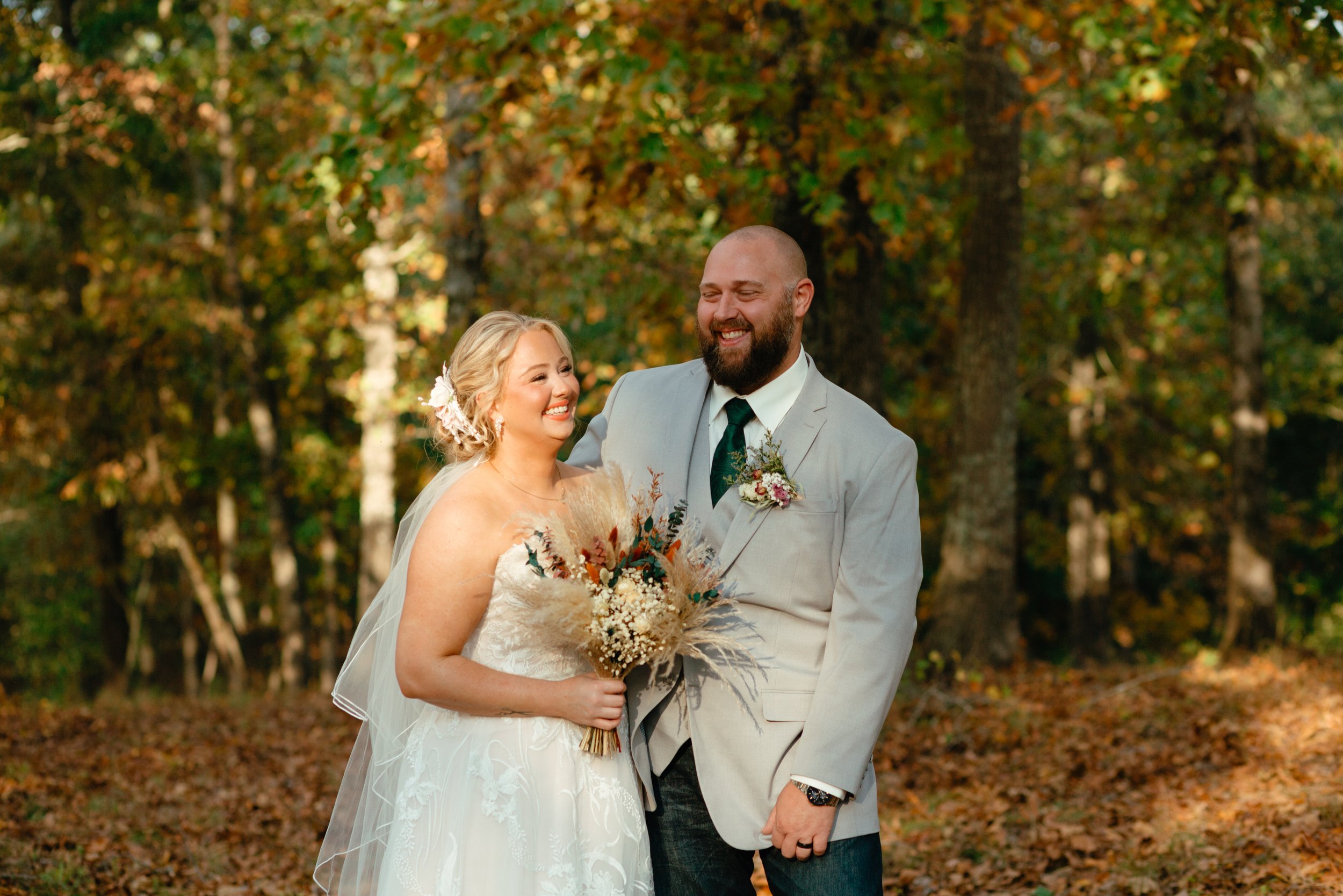 A newlywed couple standing outdoors in a forest during fall, smiling at each other. Anniston, Birmingham, Heflin, Calhoun Co, Alabama wedding photographer