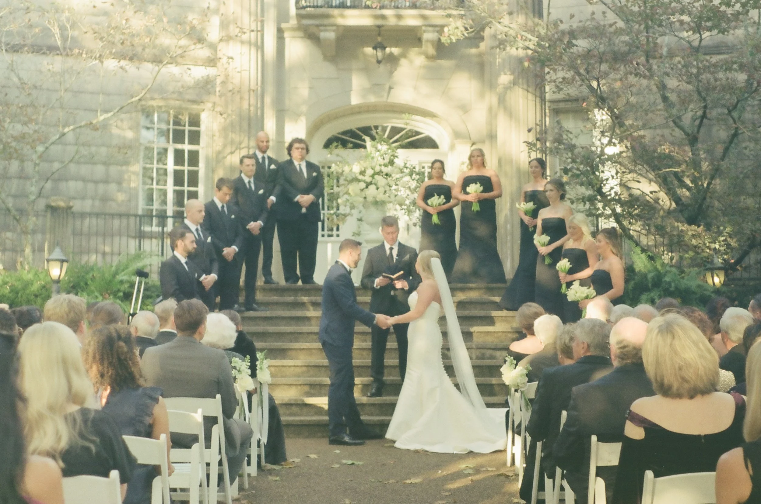 A wedding ceremony taking place outdoors on the steps of a large house or mansion. The bride and groom are holding hands and facing each other, with an officiant reading from a book. There are eight bridesmaids and groomsmen standing behind them on t
