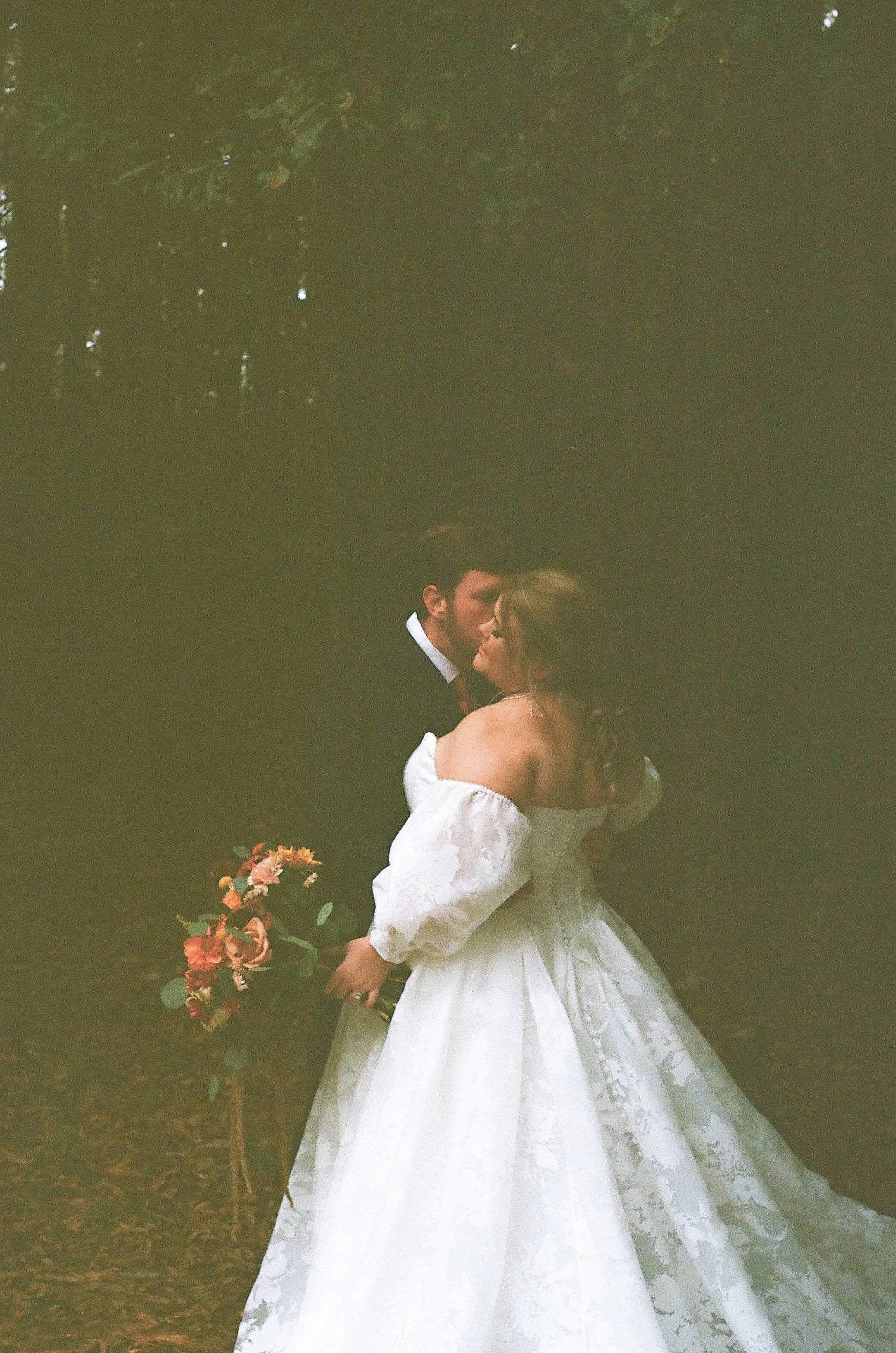 A bride and groom are sharing a kiss outdoors, with the bride holding a bouquet of flowers. The bride is wearing a white wedding gown with off-the-shoulder sleeves, and the groom is dressed in a dark suit.