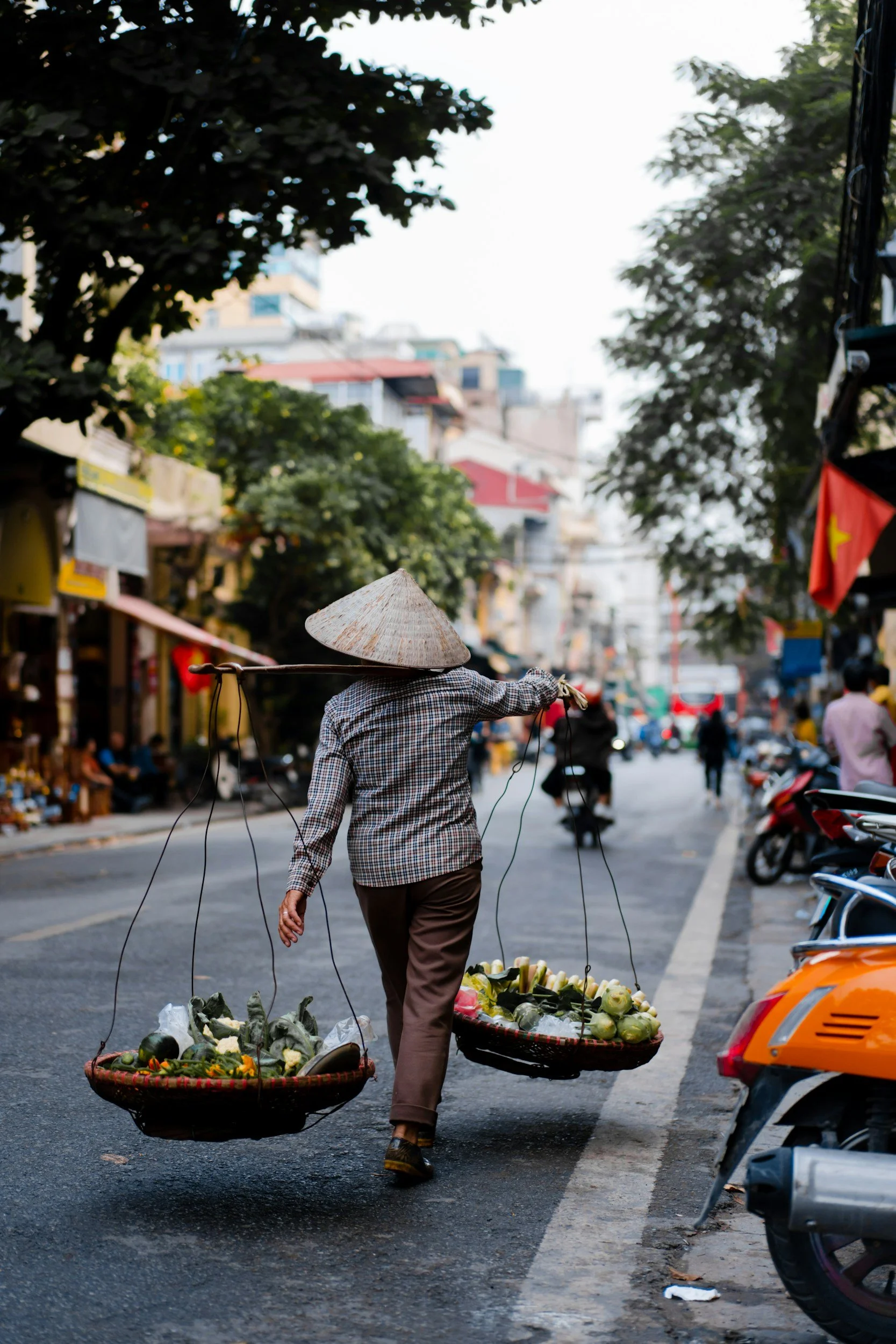 Man with traditional hat walking down street in Vietnam carrying two large baskets with fruit held on his shoulders in Vietnam with scooters in the background.