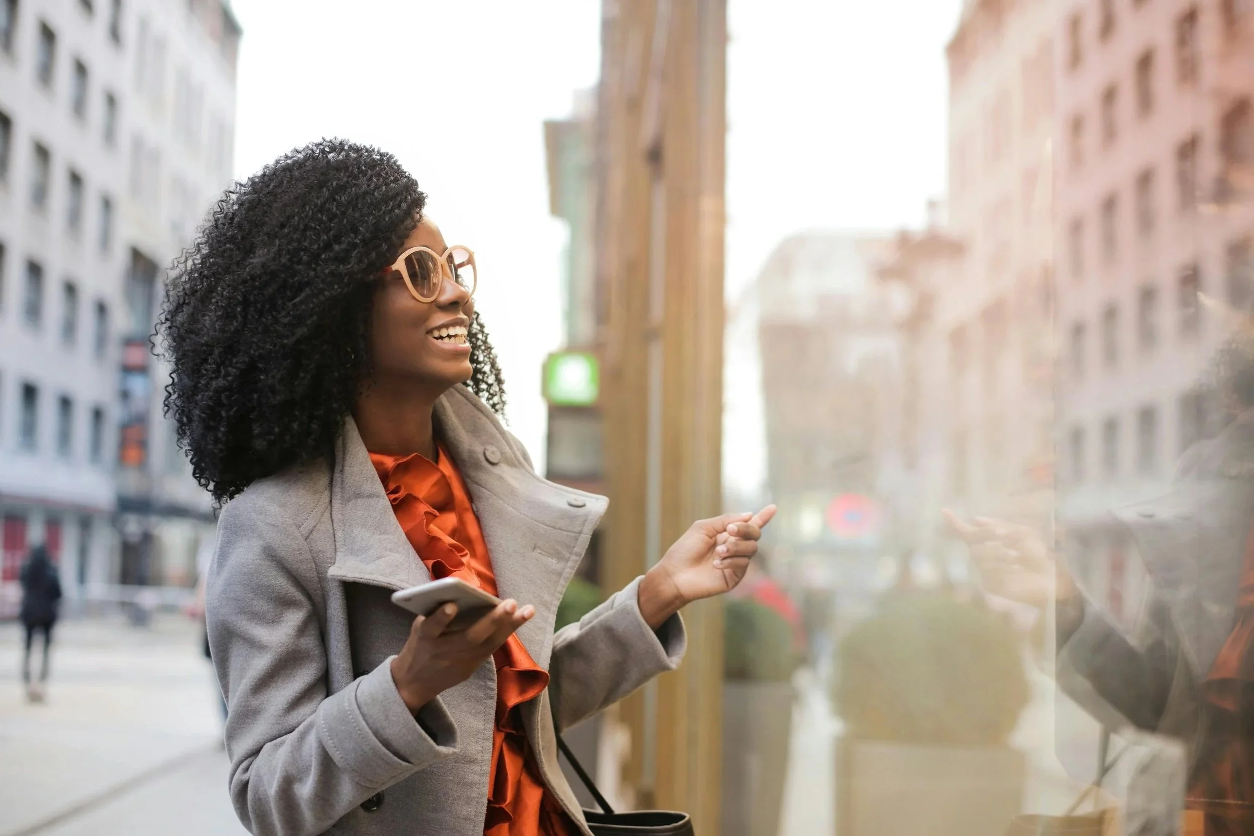 An African American woman smiling and pointing while holding a smartphone, walking on a city street with buildings in the background.
