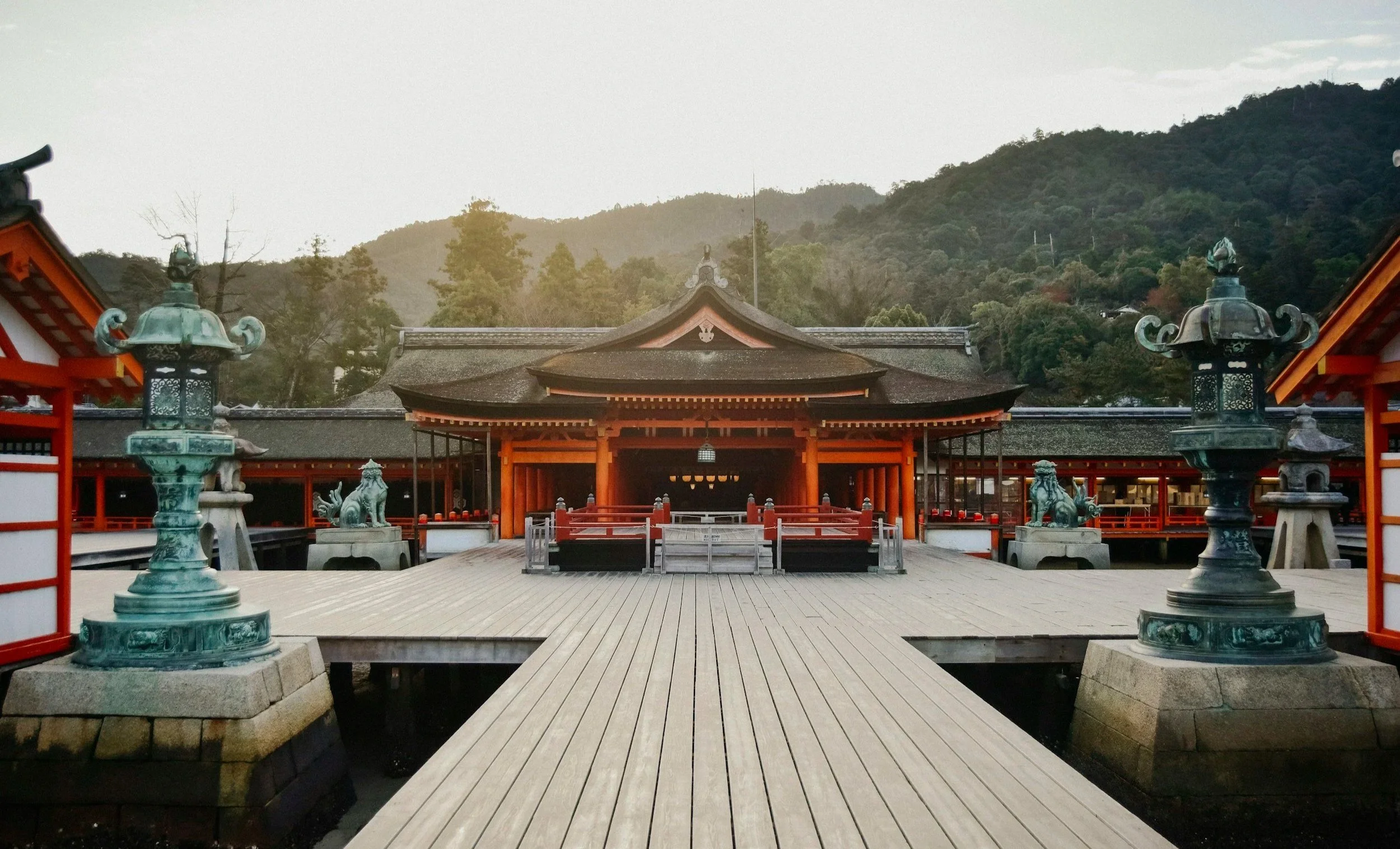 a red traditional japanese temple at the end of a wooden walking path with one teal pillar on each side and green mountains in the background
