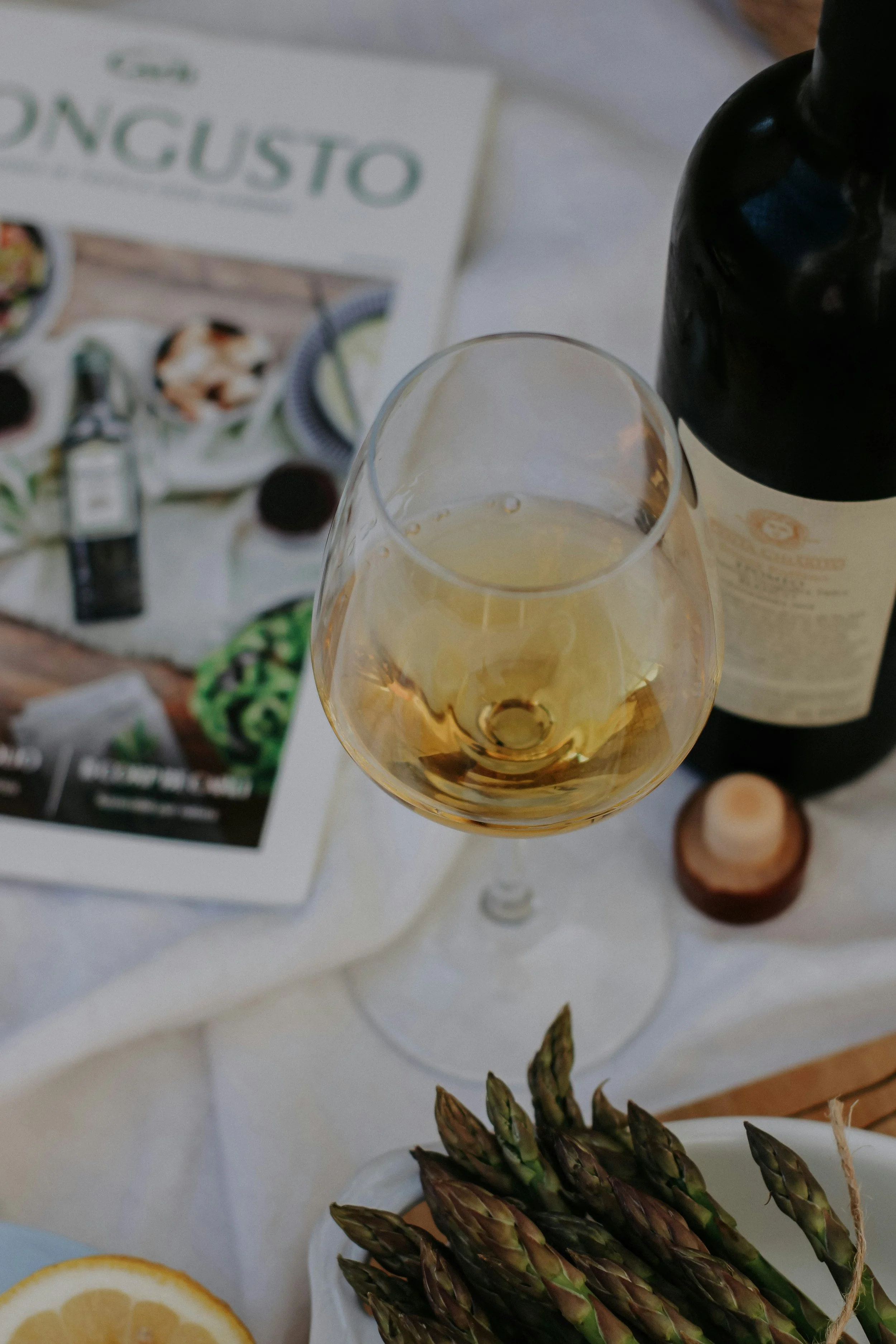 Picture of a glass of white wine, wine bottle, magazine, lemon and asparagus laid out casually on a table in Italy