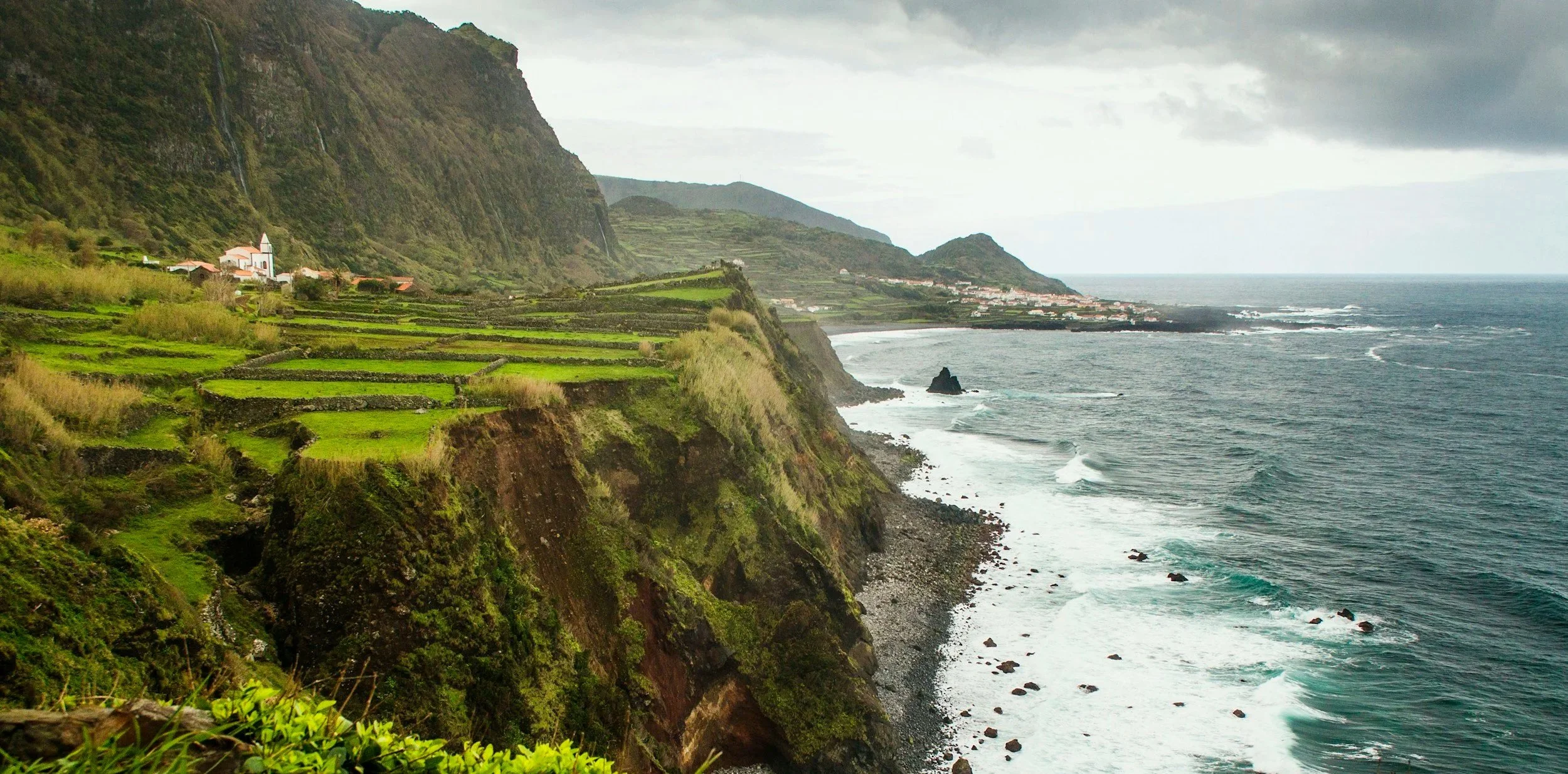Green fields sharply dropping to blue grey waters. Tall vertical mountains and a small town in the back.