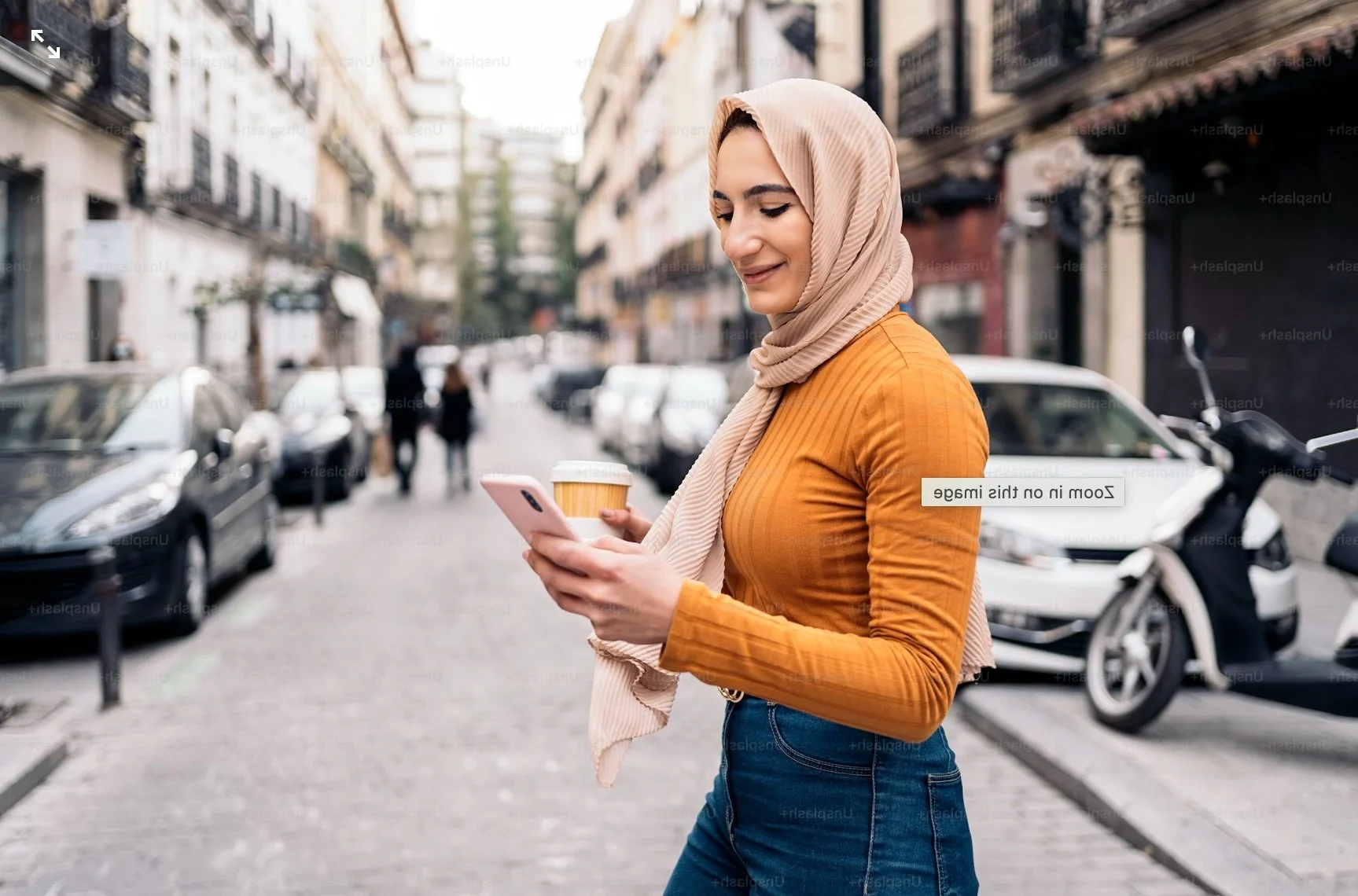 A woman wearing a beige hijab and an orange long-sleeve shirt walks on a city sidewalk, looking at her phone and holding a takeaway coffee cup.