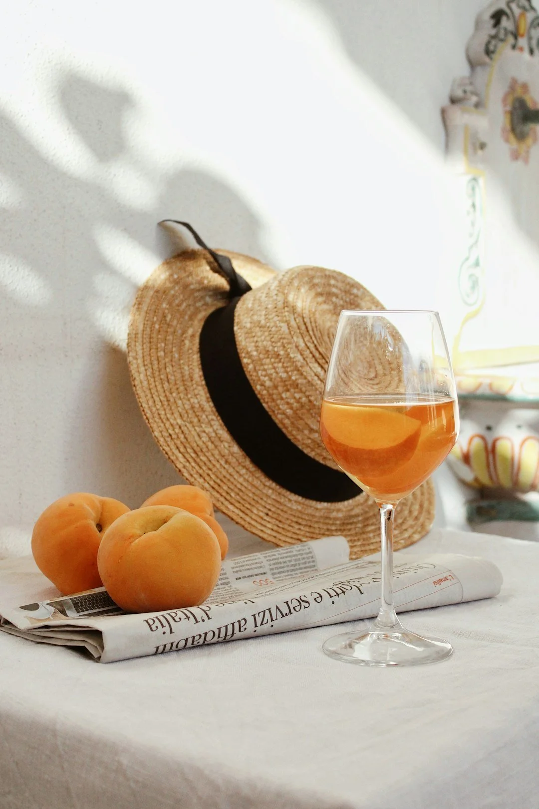 glass of orange wine on a white table with a folded italian newspaper, 3 peaches ad a straw hat with black ribbon.