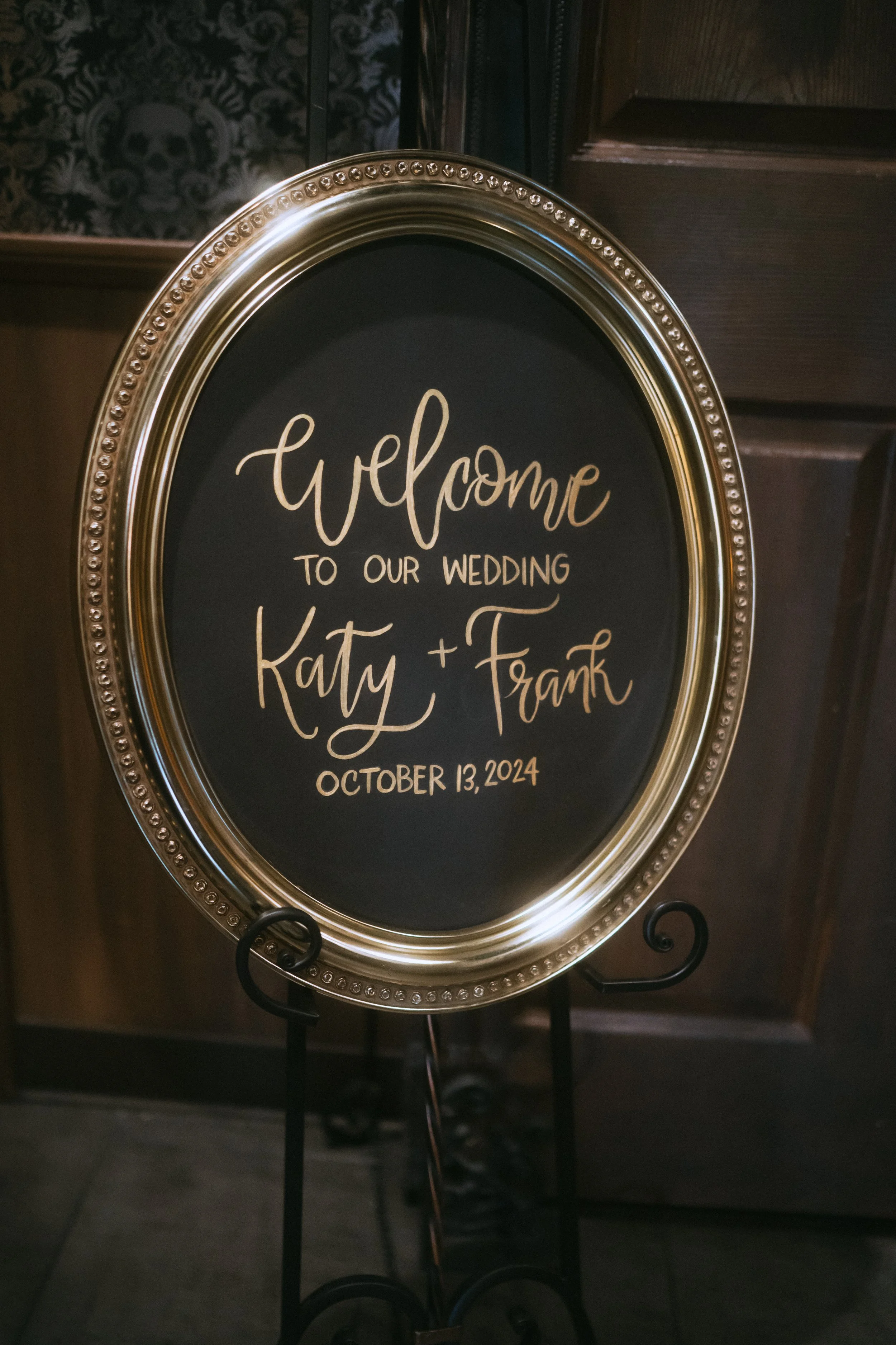 Round black and gold wedding welcome sign reading “Katy + Frank, October 13, 2024” displayed on an easel