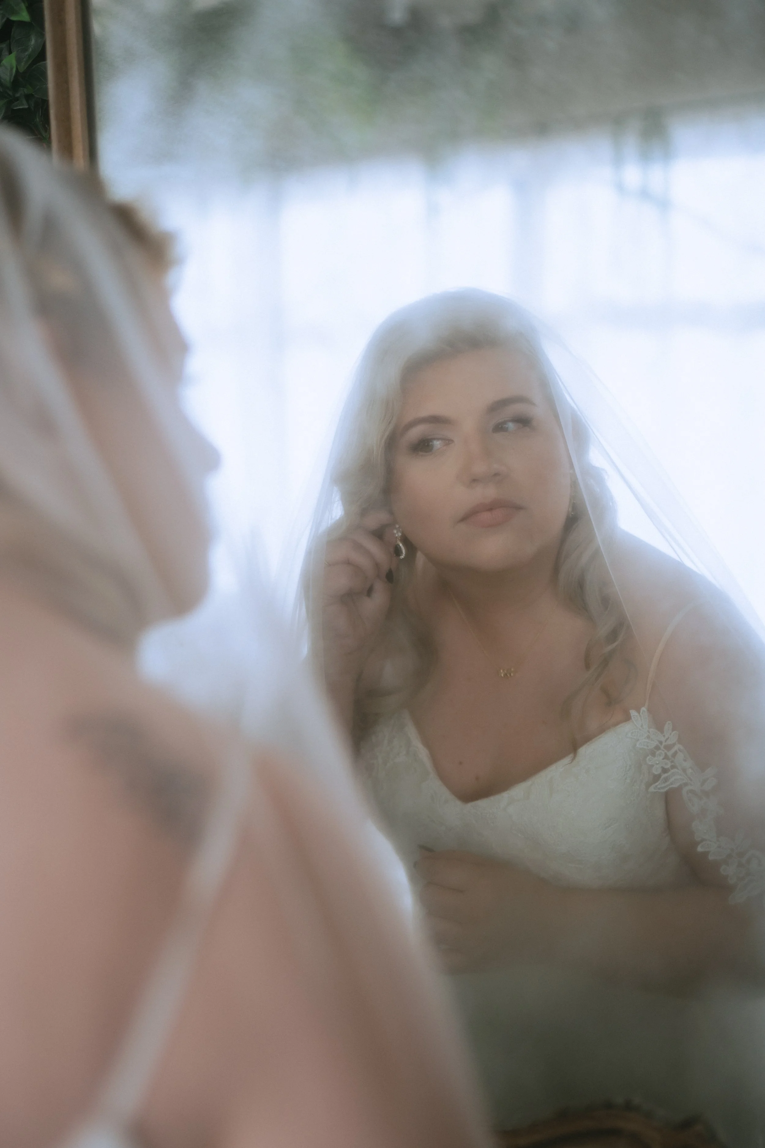 Bride getting ready on her wedding morning, adjusting her earring in the mirror while wearing a veil and lace gown