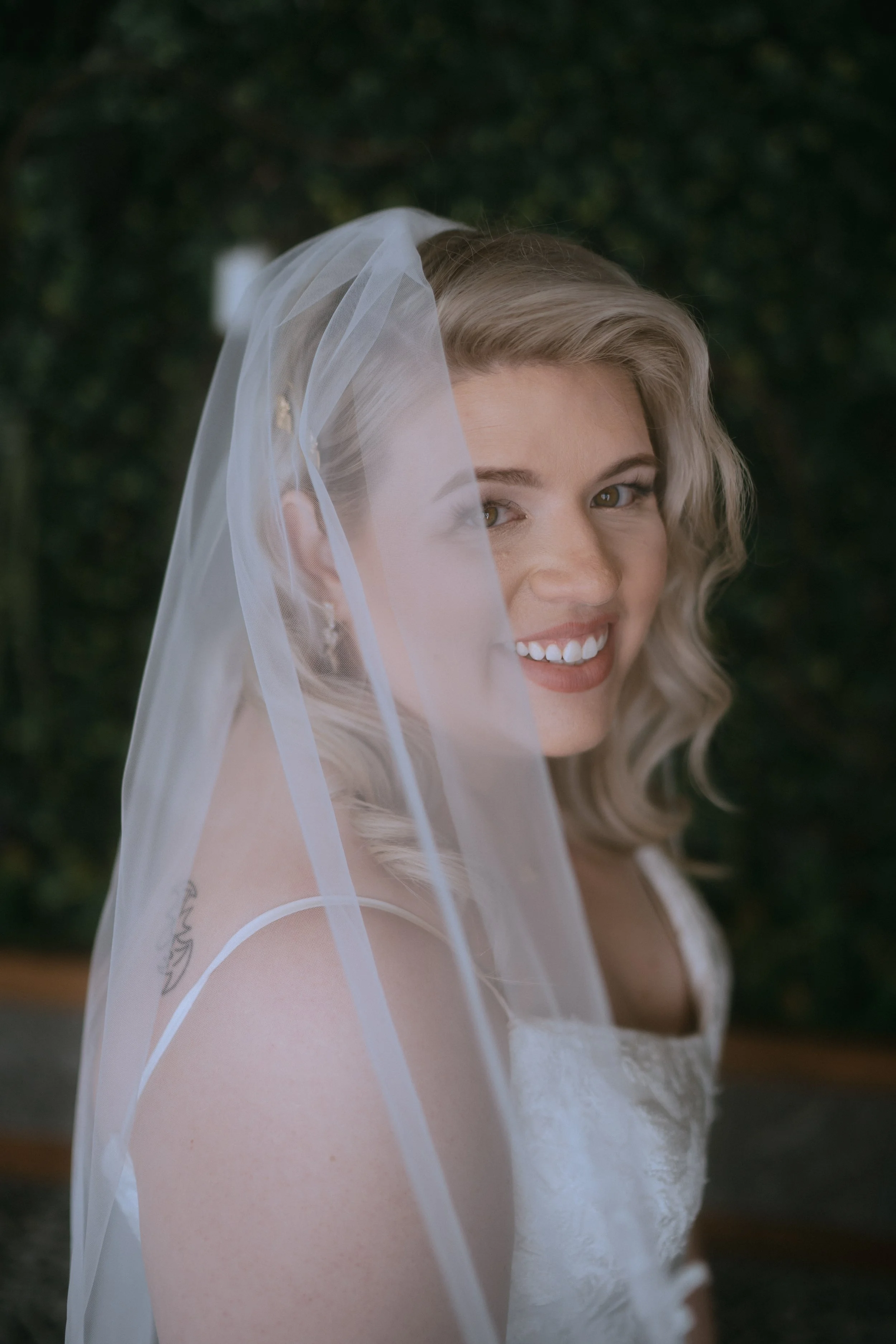 Bride smiling in soft natural light with veil draped over her face during wedding day portraits