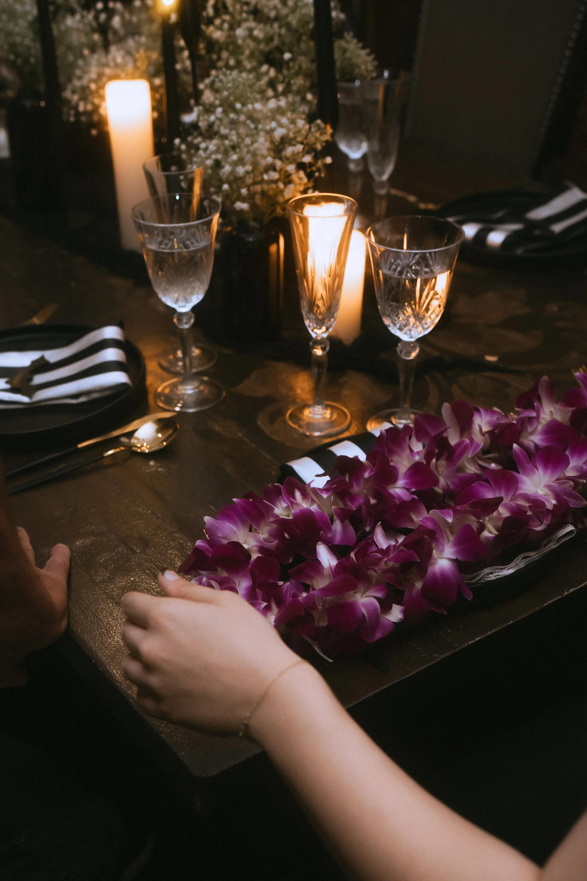Moody wedding reception table with candlelight, crystal glassware, striped napkins, and purple orchid lei detail