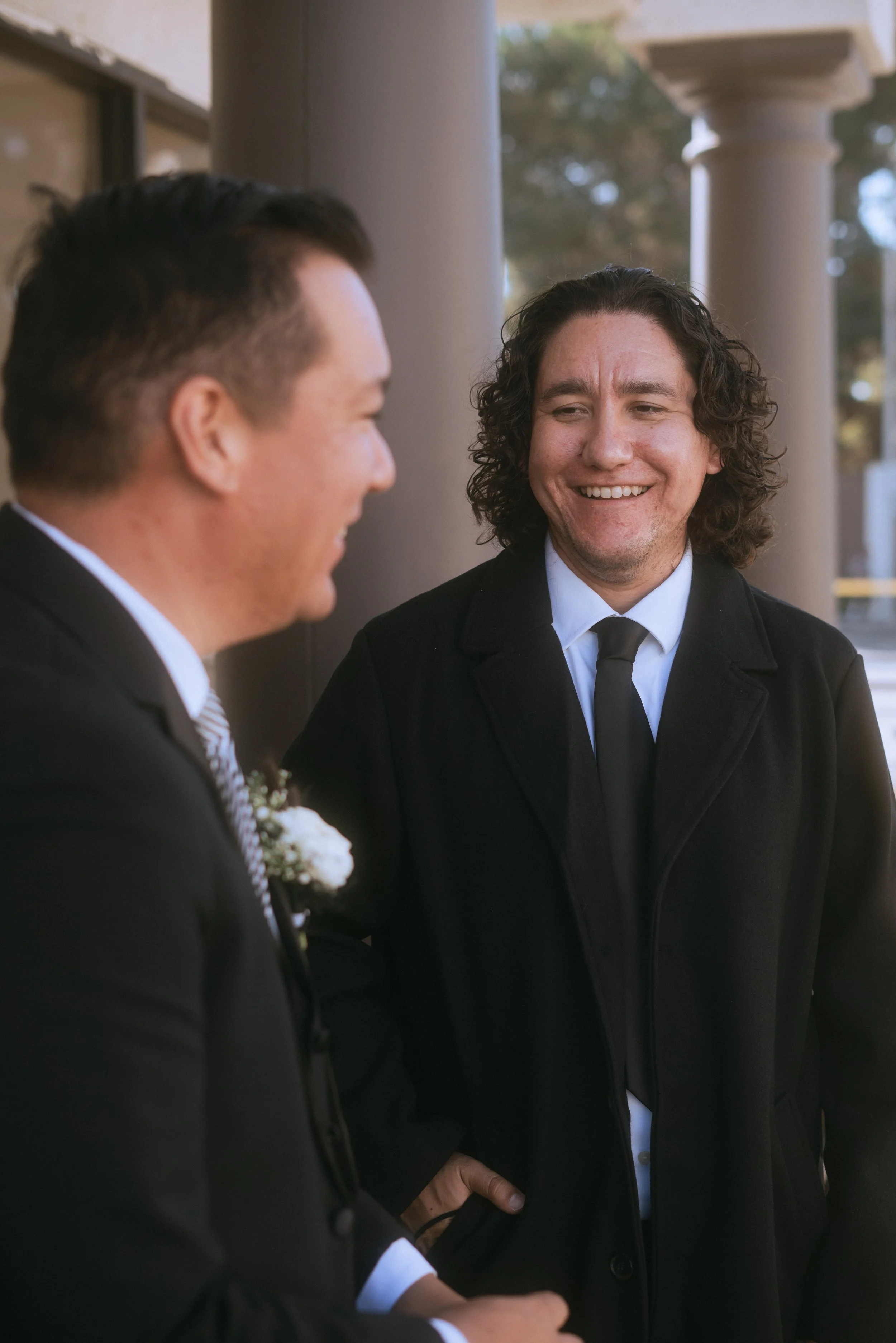 Groom in black suit smiling and talking with another man outside before the wedding ceremony