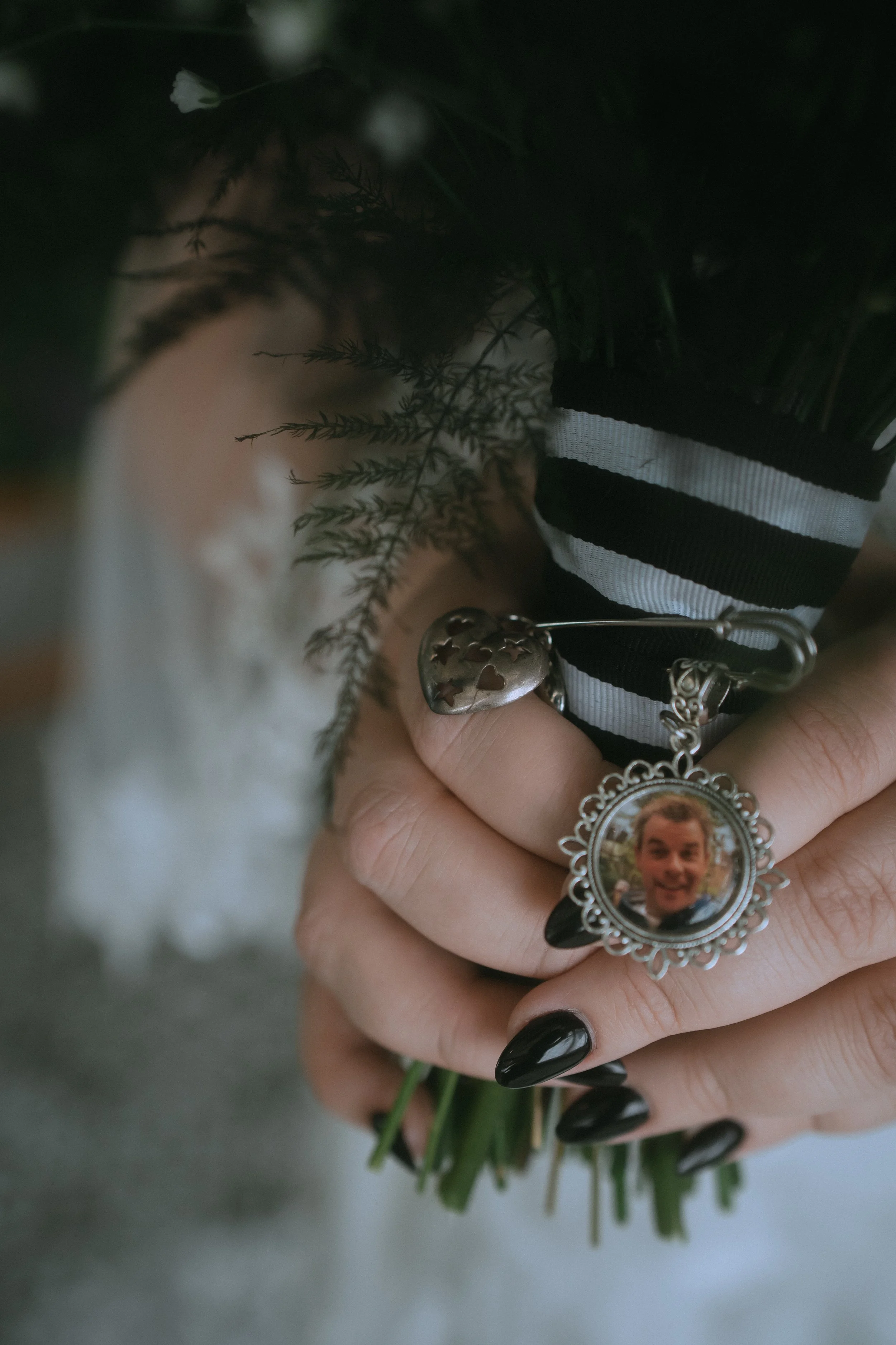 Close-up of bride holding bouquet with a vintage locket photo attached, black ribbon wrap, and dark manicure detail