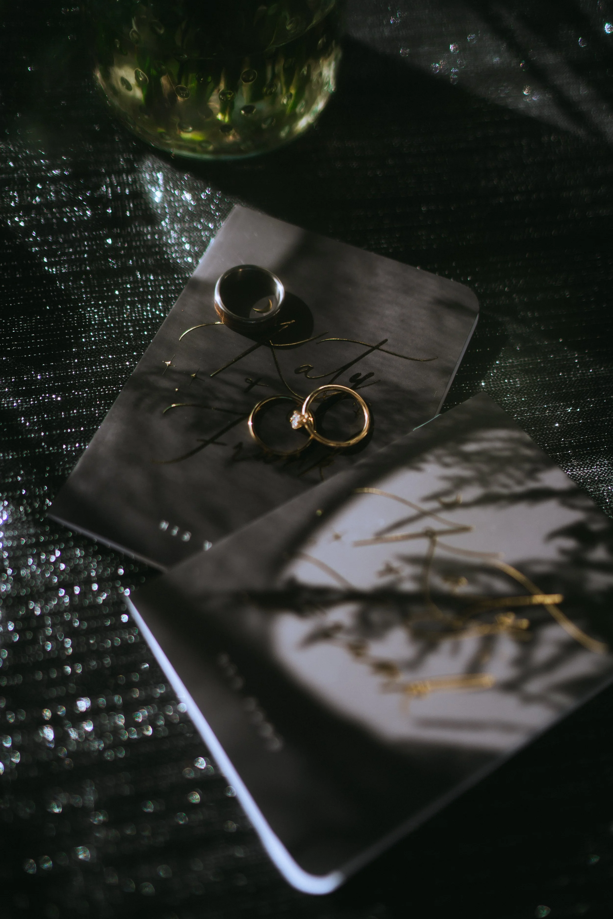 Wedding rings styled on invitation suite in a dark romantic flat lay with textured background and glass detail