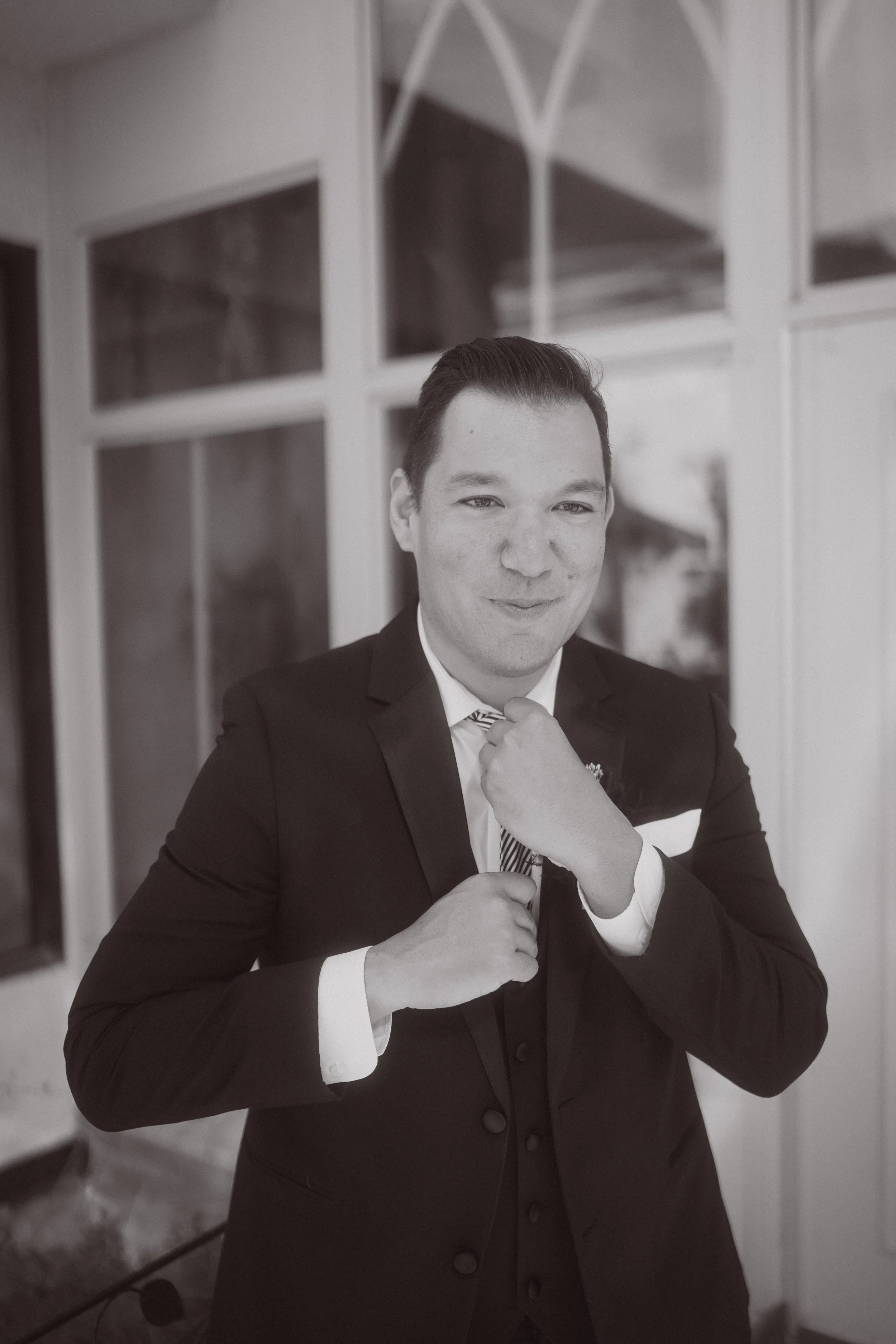 Groom adjusting his tie in a black and white portrait during wedding getting ready moments indoors