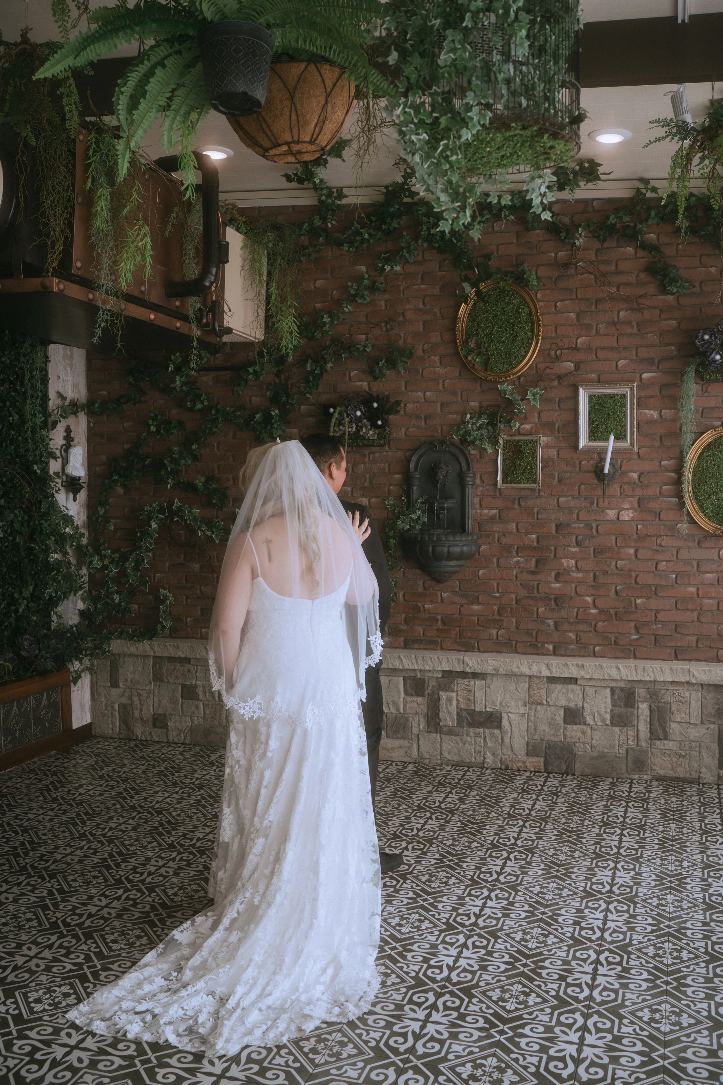 Wide shot of bride and groom during first look inside venue with greenery, brick wall, and patterned tile floor