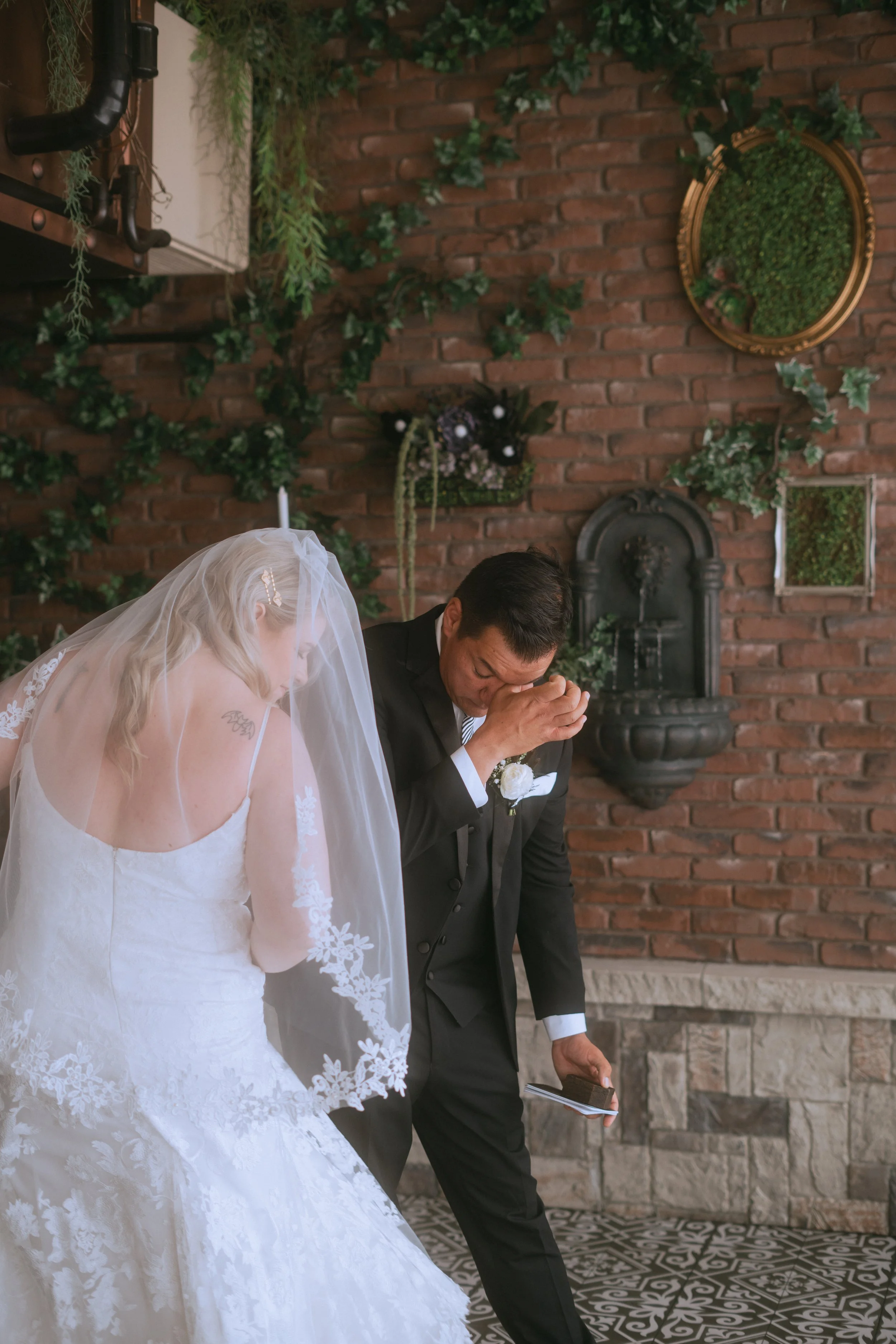 Groom wiping away tears during emotional first look with bride in veil inside indoor wedding venue with brick wall and greenery