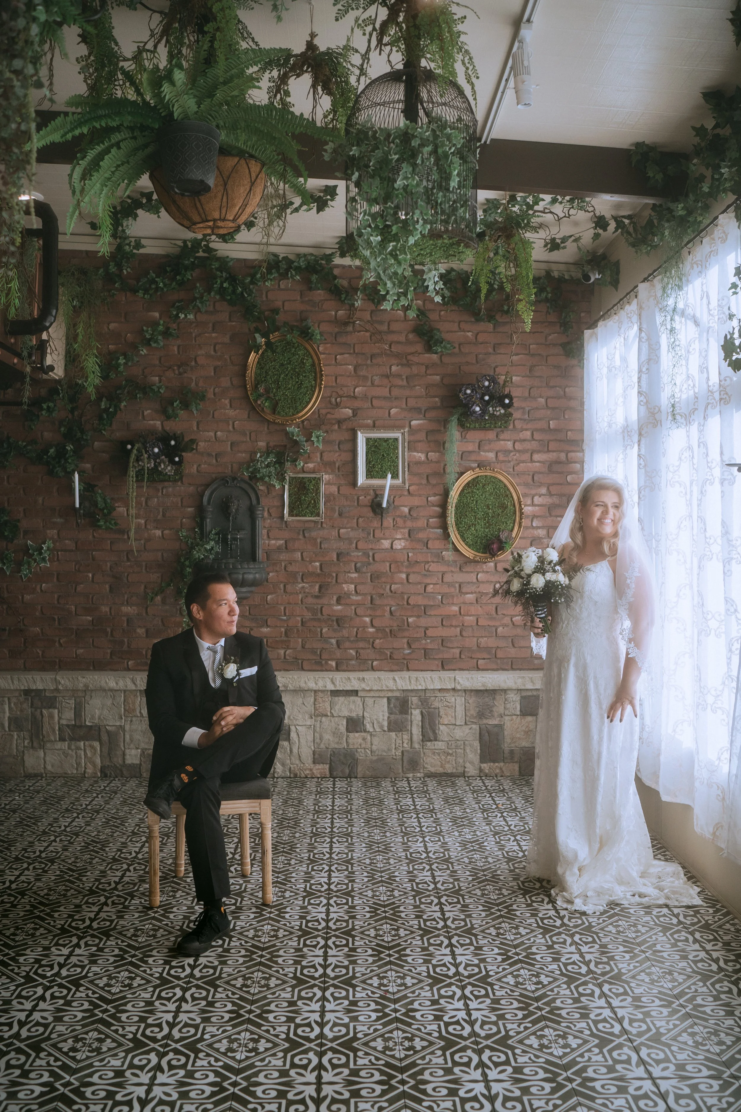 Bride holding bouquet near window while groom sits across the room inside decorated indoor wedding venue with greenery and patterned tile floor