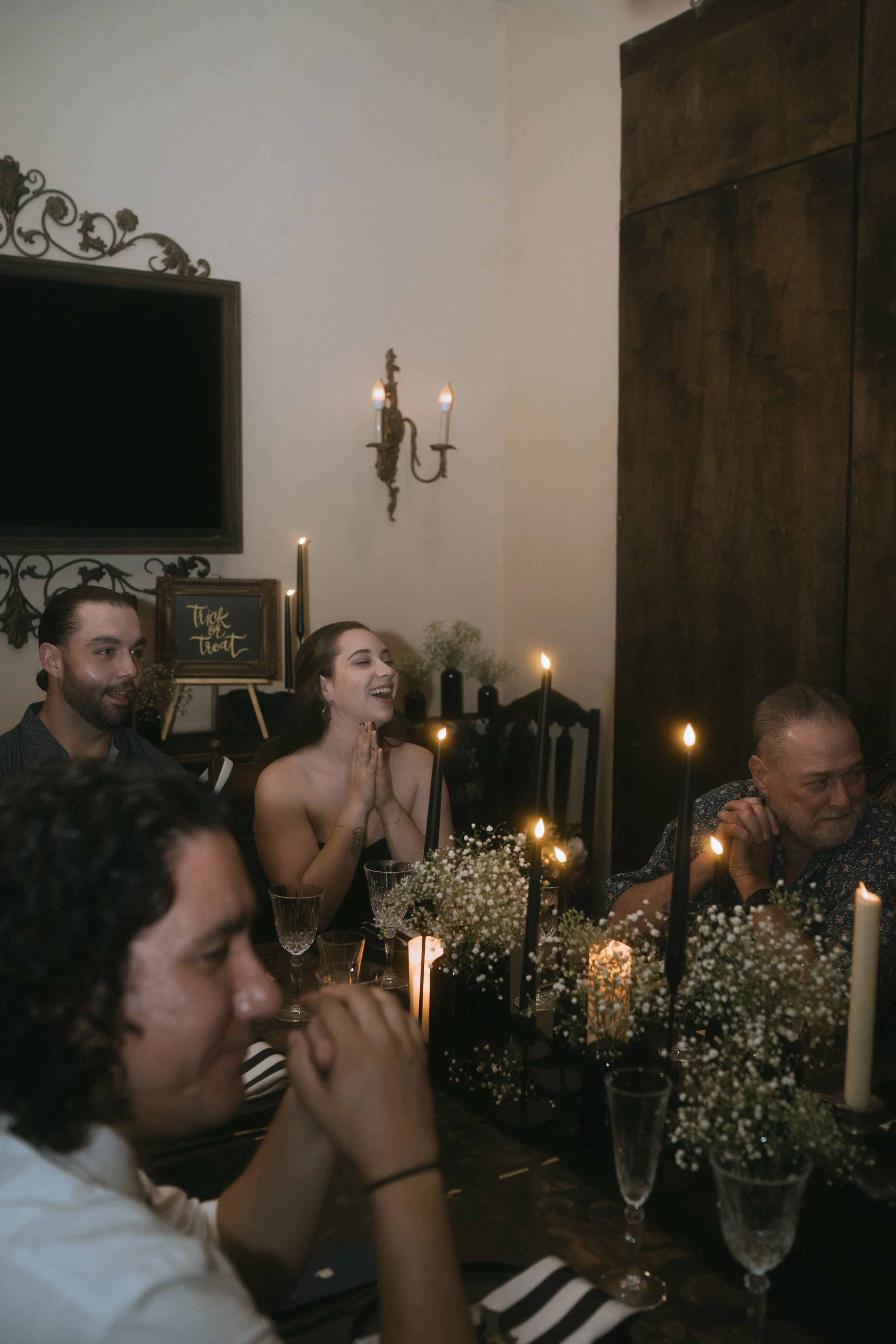 Wedding guests laughing and talking at candlelit reception table with floral centerpieces and dark romantic decor