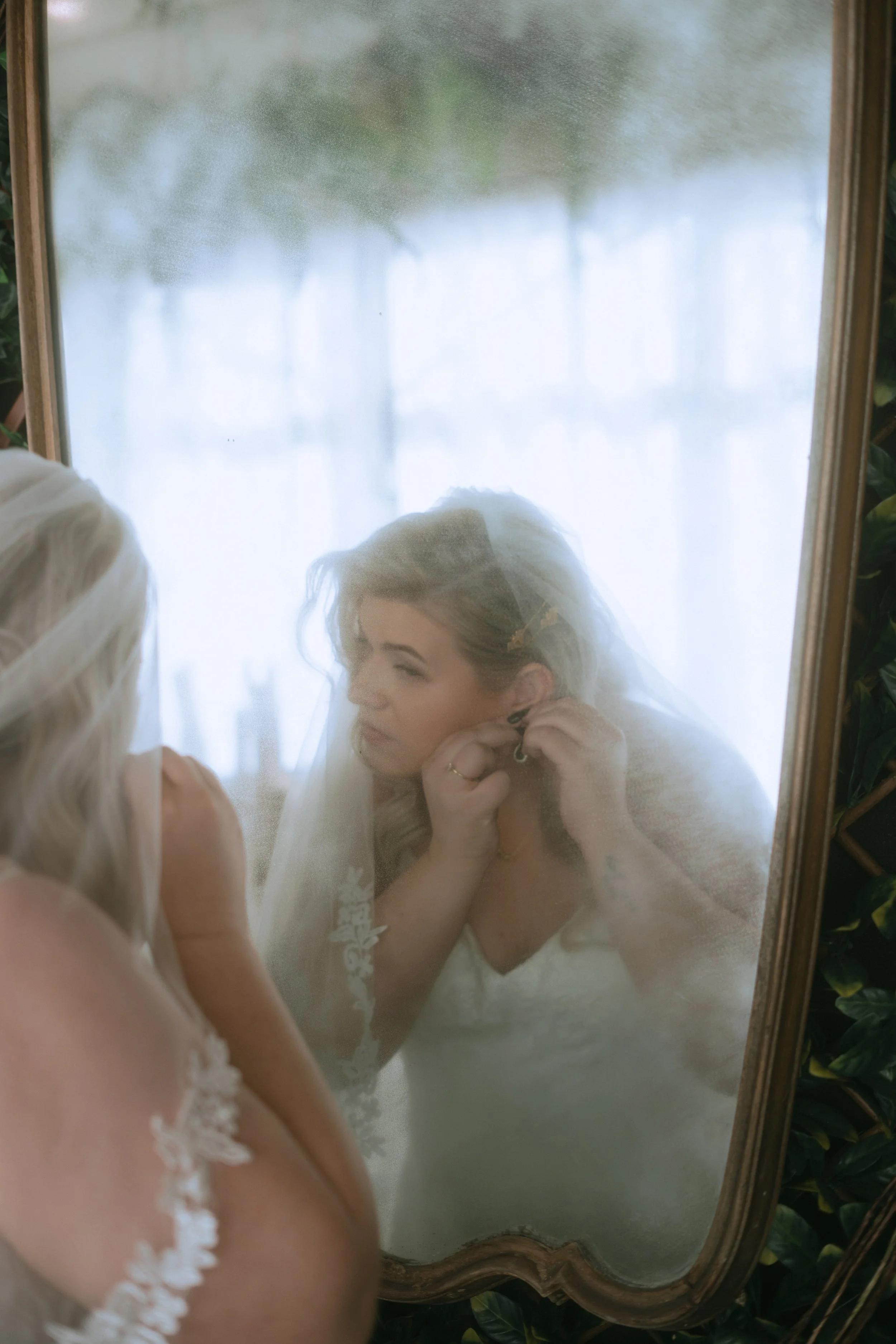 Bride putting on earrings in front of a mirror during wedding morning getting ready with soft natural light